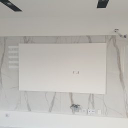 ŁUKASZ CYZ Budo - Solid - Ściana z płytami imitującymi marmur, z centralnie umieszczonym białym panelem na telewizor, widoczne gniazdka elektryczne i puszka instalacyjna w górnym rogu, sufit z czarnymi reflektorami.