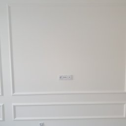 ŁUKASZ CYZ Budo - Solid - Biała ściana z dekoracyjnymi listwami, gniazdkami i widocznym oknem z żaluzjami po lewej stronie.