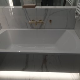 ŁUKASZ CYZ Budo - Solid - Biała wanna obudowana marmurowymi płytami, złota bateria ścienna, grzejnik drabinkowy w łazience.