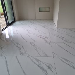 ŁUKASZ CYZ Budo - Solid - Błyszczące, białe płytki podłogowe imitujące marmur w nowym, jasnym pomieszczeniu z widocznym oknem i przygotowaną wnęką w ścianie.