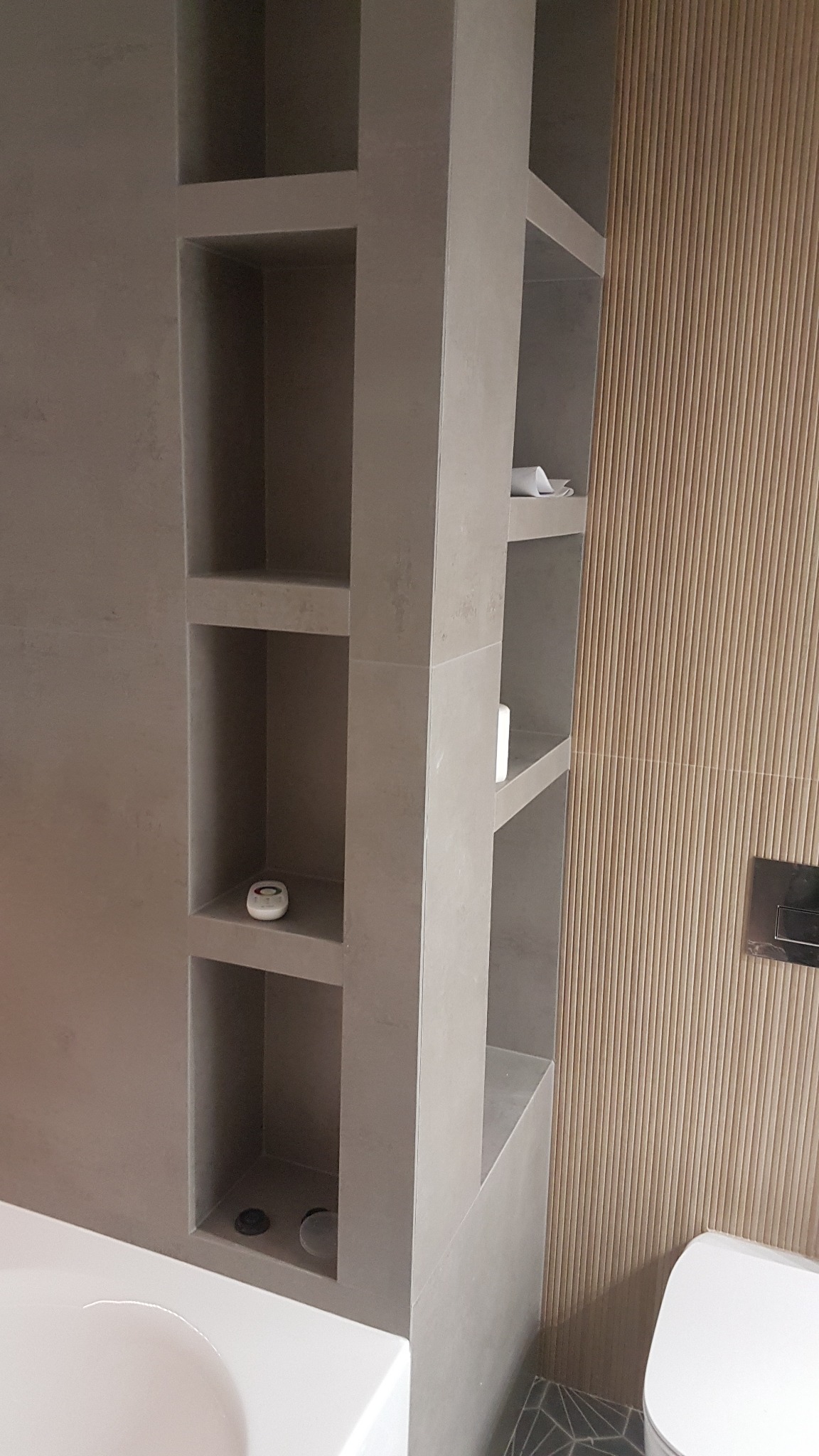 Półki wnękowe z szarej płytki w łazience, na półkach kosmetyki i ręczniki, obok ściana z drewnopodobnych paneli pionowych, widoczny fragment wanny i toalety.
