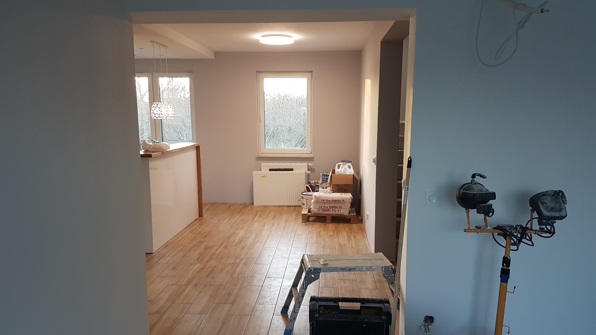 Wnętrze w trakcie prac wykończeniowych: drewnopodobna podłoga, świeżo pomalowane szare ściany, widoczne worki z zaprawą, drabina malarska i lampa budowlana na statywie.