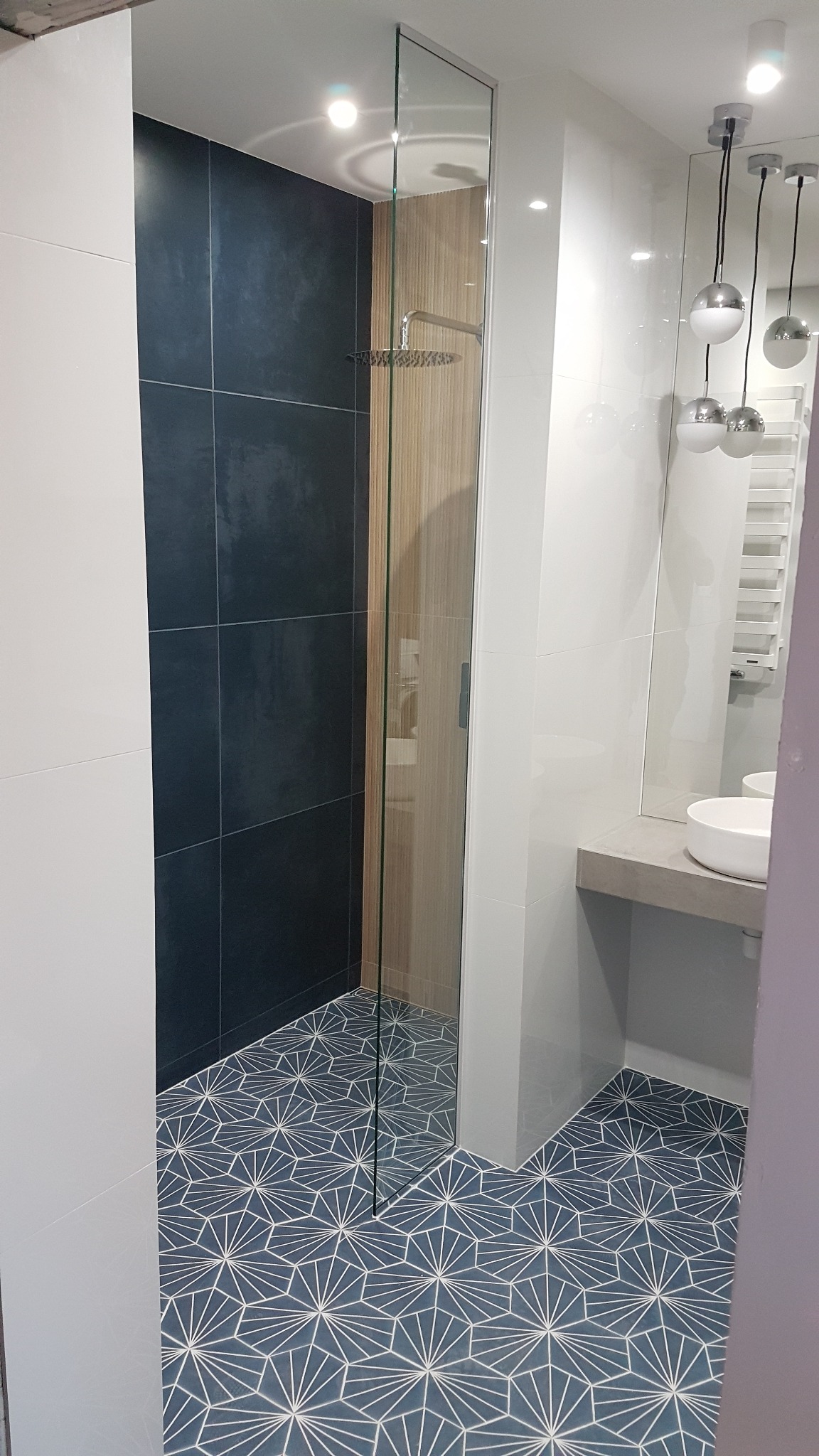 Nowoczesna łazienka z kabiną prysznicową typu walk-in, ciemnymi płytkami pod prysznicem, wzorzystą podłogą w odcieniach niebieskiego i białego oraz wiszącą lampą z trzema kulami.
