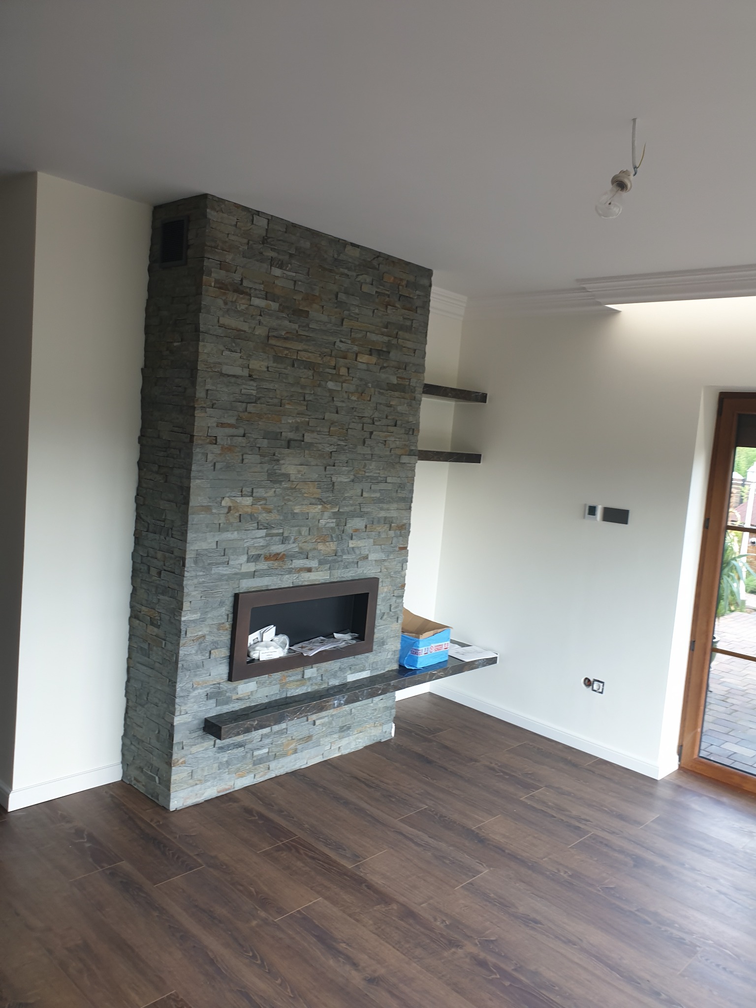 Wykończony kominek obłożony szarym kamieniem dekoracyjnym, z drewnianą obudową paleniska, na podłodze panele imitujące ciemne drewno, widoczne półki na ścianie i niedokończone oświetlenie sufitowe.