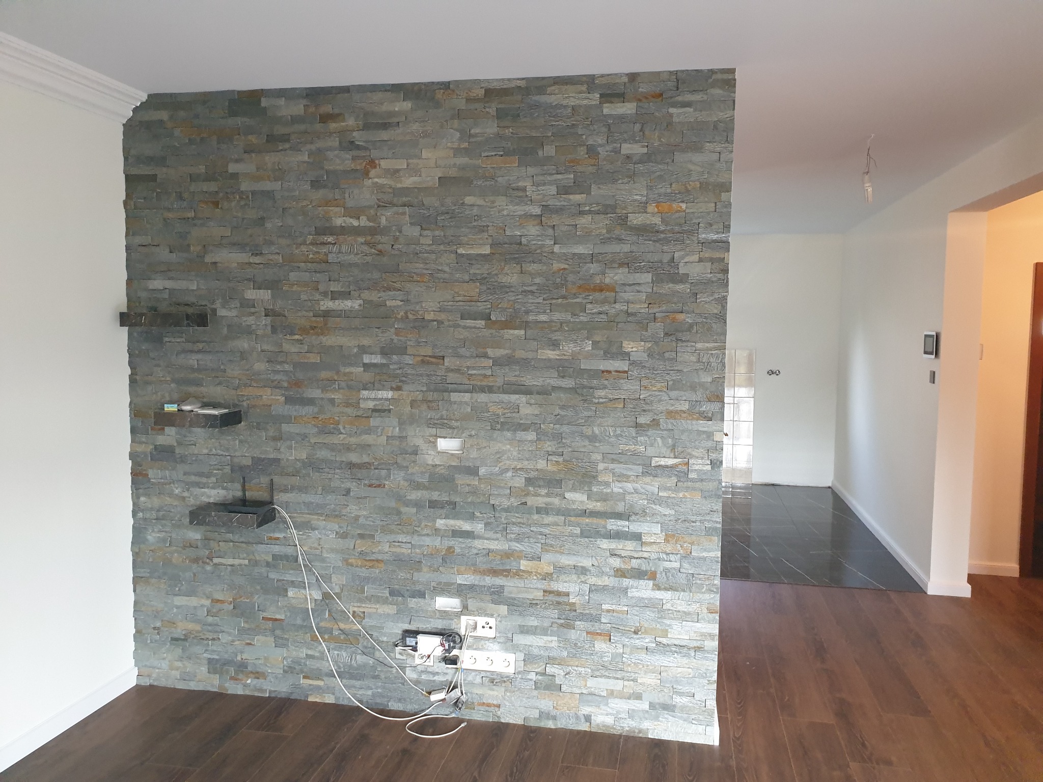 Ściana działowa wyłożona szarą okładziną kamienną w salonie, z widocznymi gniazdkami elektrycznymi i półkami z kamienia, podłoga z paneli imitujących drewno.