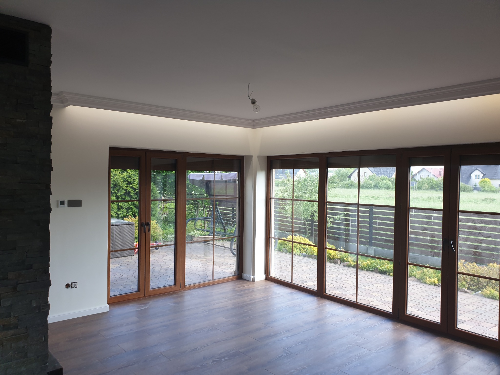Przestronne wnętrze z dużymi oknami tarasowymi z drewnianą ramą, parkiet na podłodze, fragment ściany wykończony kamieniem dekoracyjnym, widoczna listwa sufitowa z oświetleniem LED i goła żarówka