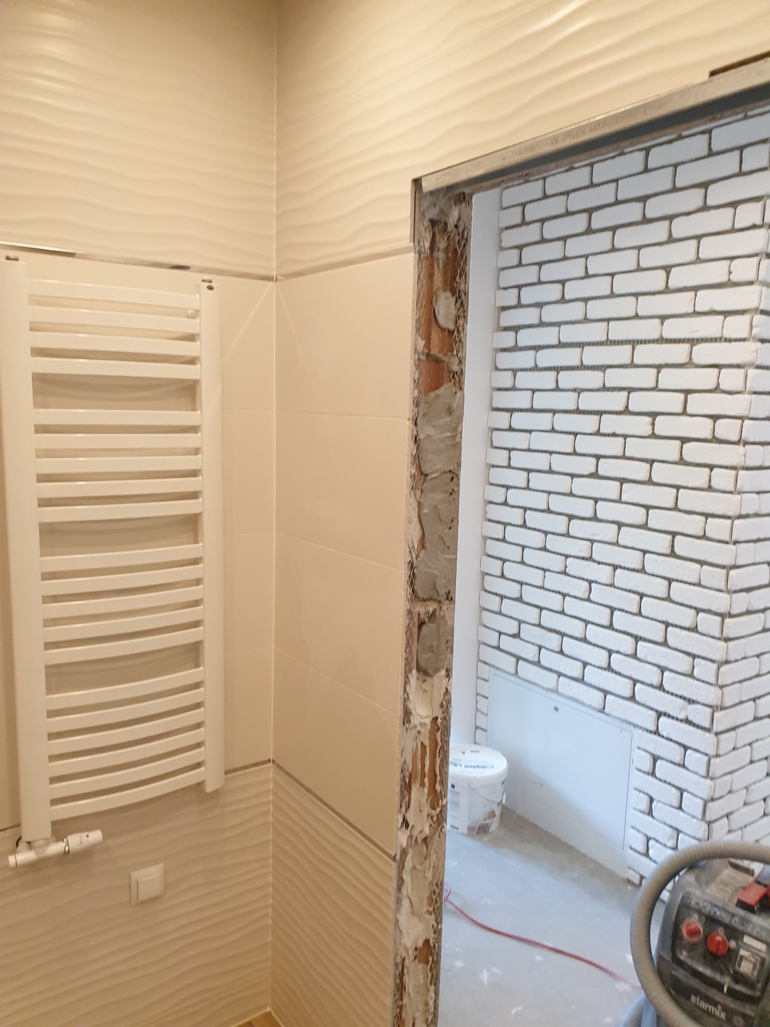 Łazienka w trakcie prac: białe płytki imitujące cegłę na ścianie, widoczne surowe wykończenie ościeżnicy, grzejnik drabinkowy na ścianie z kremowymi płytkami z falistą fakturą, odkurzacz...