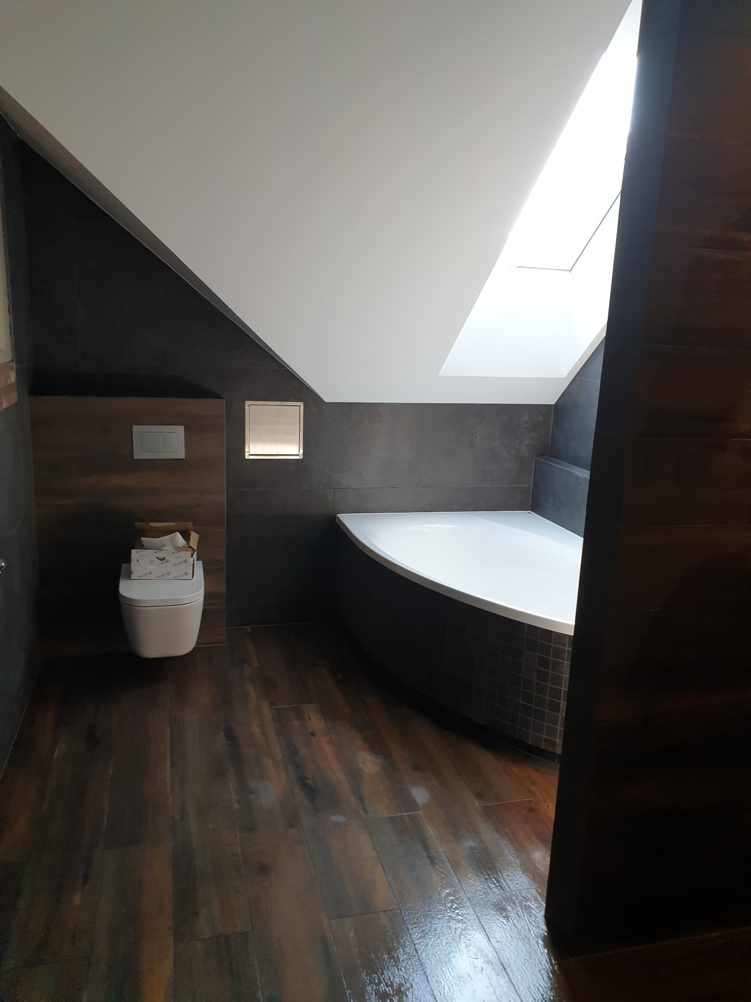 Nowoczesna łazienka na poddaszu z drewnopodobną podłogą, ciemnymi płytkami ściennymi, białą wanną z mozaiką, toaletą z pudełkiem na sedesie i oknem dachowym.