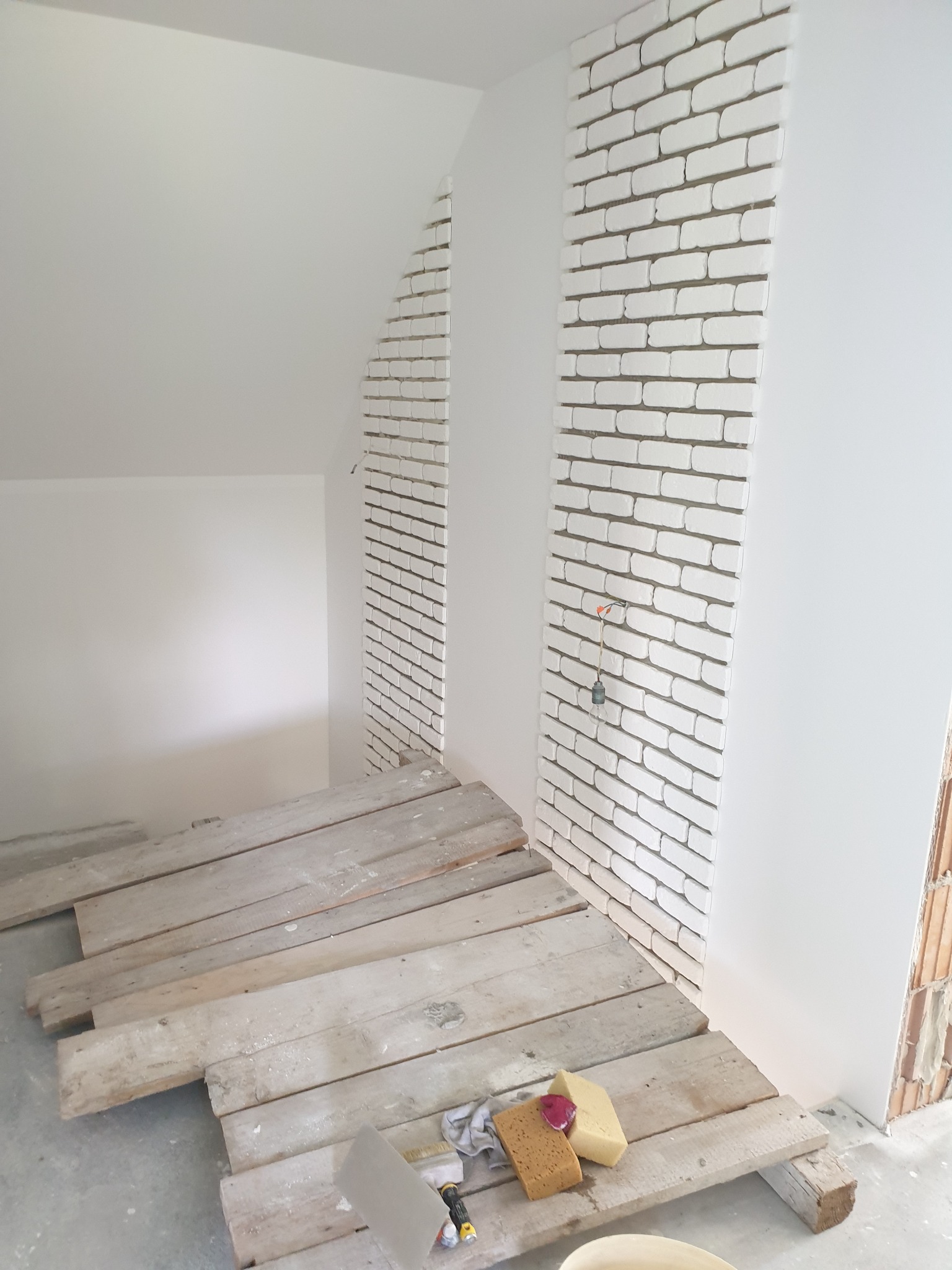 Ściana z białej cegły klinkierowej w trakcie montażu, widoczne drewniane deski zabezpieczające podłogę i akcesoria glazurnicze, surowy stan poddasza.