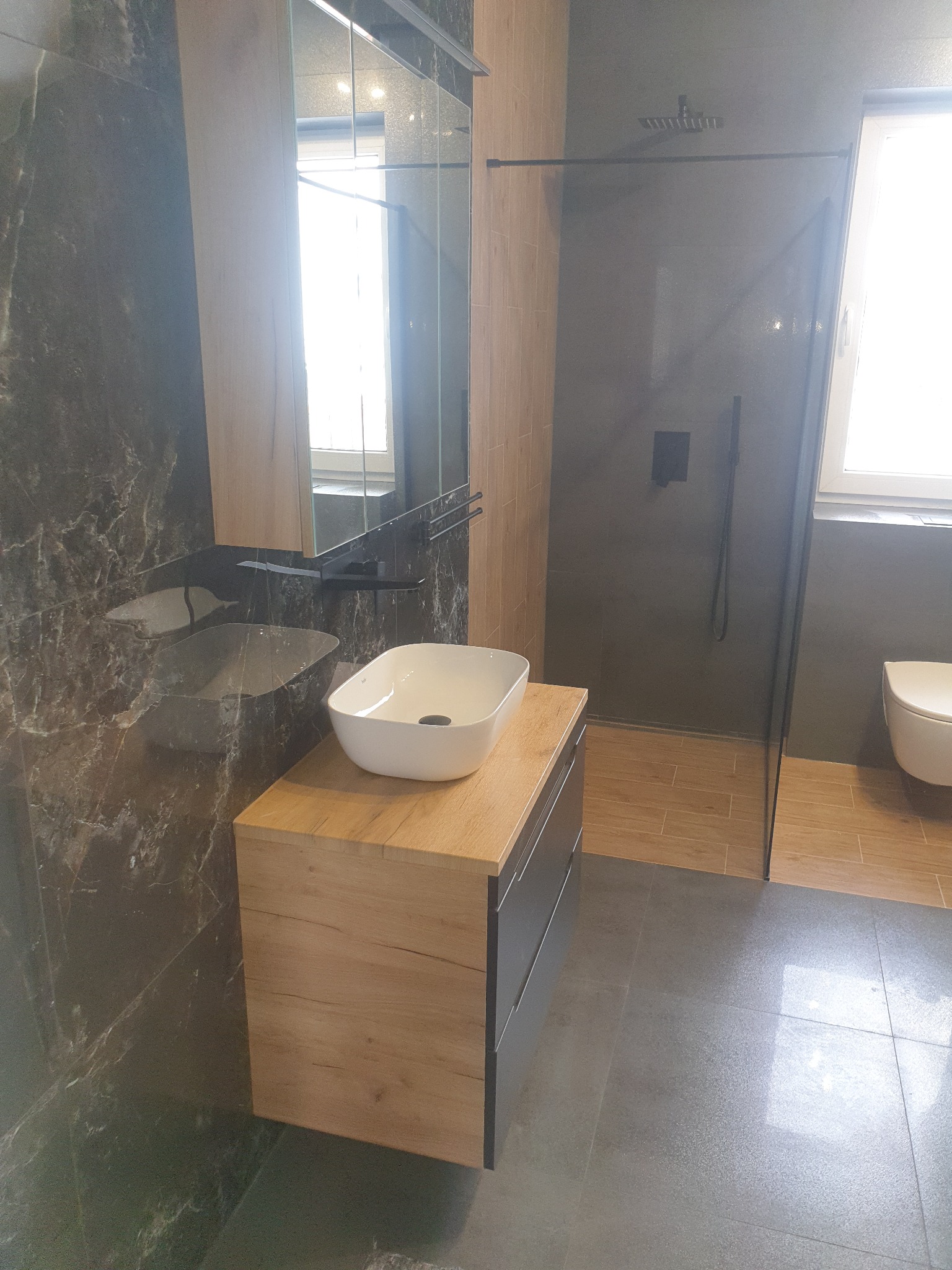 Nowoczesna łazienka z umywalką nablatową na drewnianej szafce, kabiną prysznicową typu walk-in z czarną armaturą i marmurowymi płytkami na ścianie.
