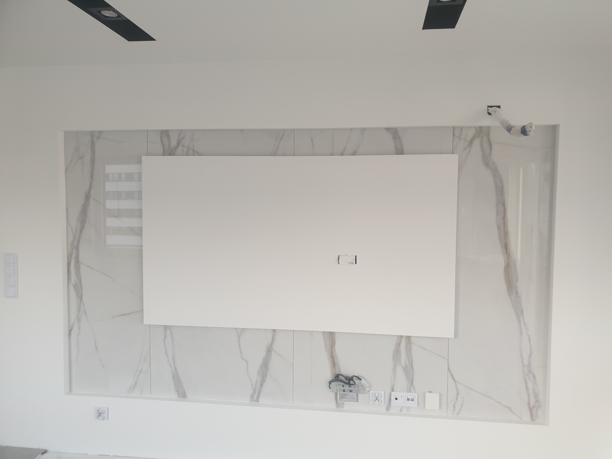 Ściana z płytami imitującymi marmur, z centralnie umieszczonym białym panelem na telewizor, widoczne gniazdka elektryczne i puszka instalacyjna w górnym rogu, sufit z czarnymi reflektorami.