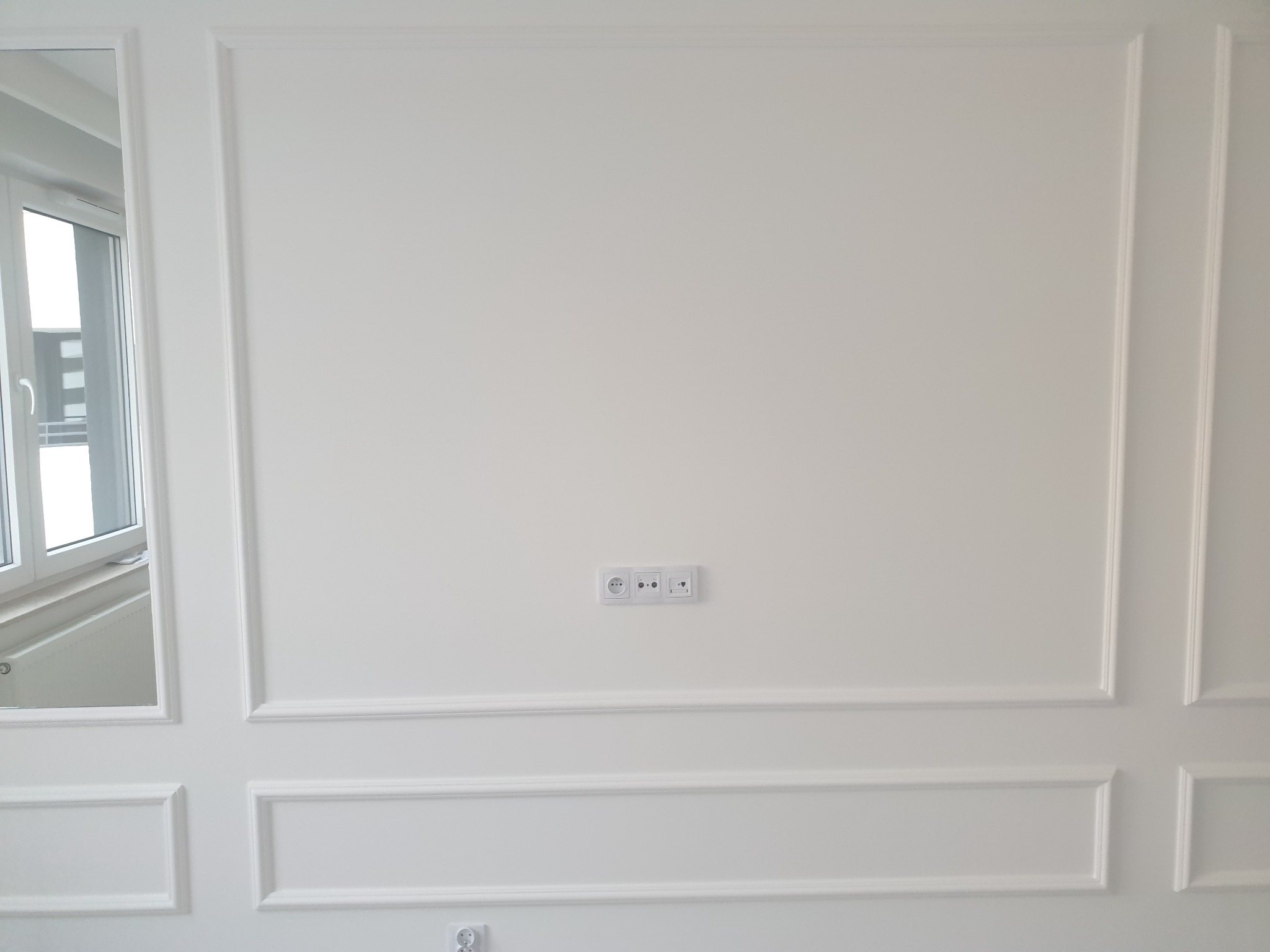 Biała ściana z dekoracyjnymi listwami, gniazdkami i widocznym oknem z żaluzjami po lewej stronie.