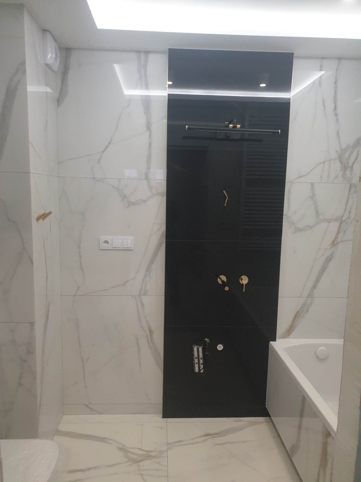 Nowoczesna łazienka z białymi marmurkowymi płytkami i czarną ścianą prysznicową z złotymi elementami armatury, oświetleniem LED i częściowo widoczną białą wanną.