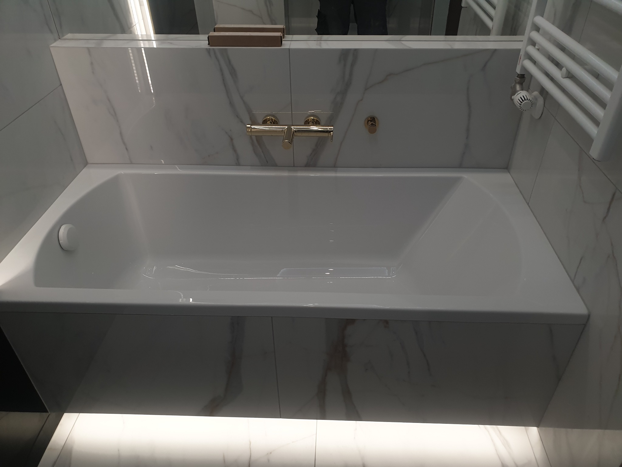 Biała wanna obudowana marmurowymi płytami, złota bateria ścienna, grzejnik drabinkowy w łazience.