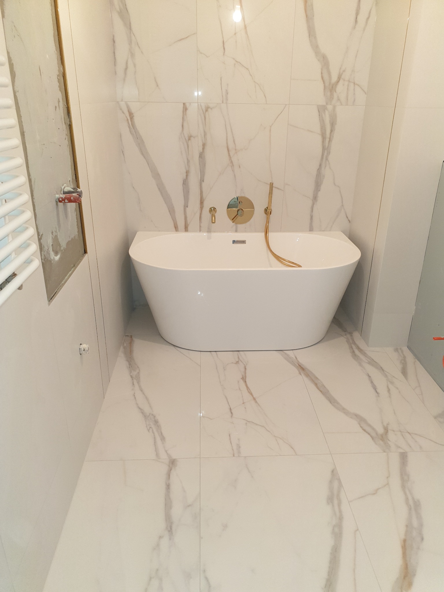 Biała, wolnostojąca wanna z złotą armaturą na tle marmurowych płytek, częściowo wykończona ściana z grzejnikiem drabinkowym.