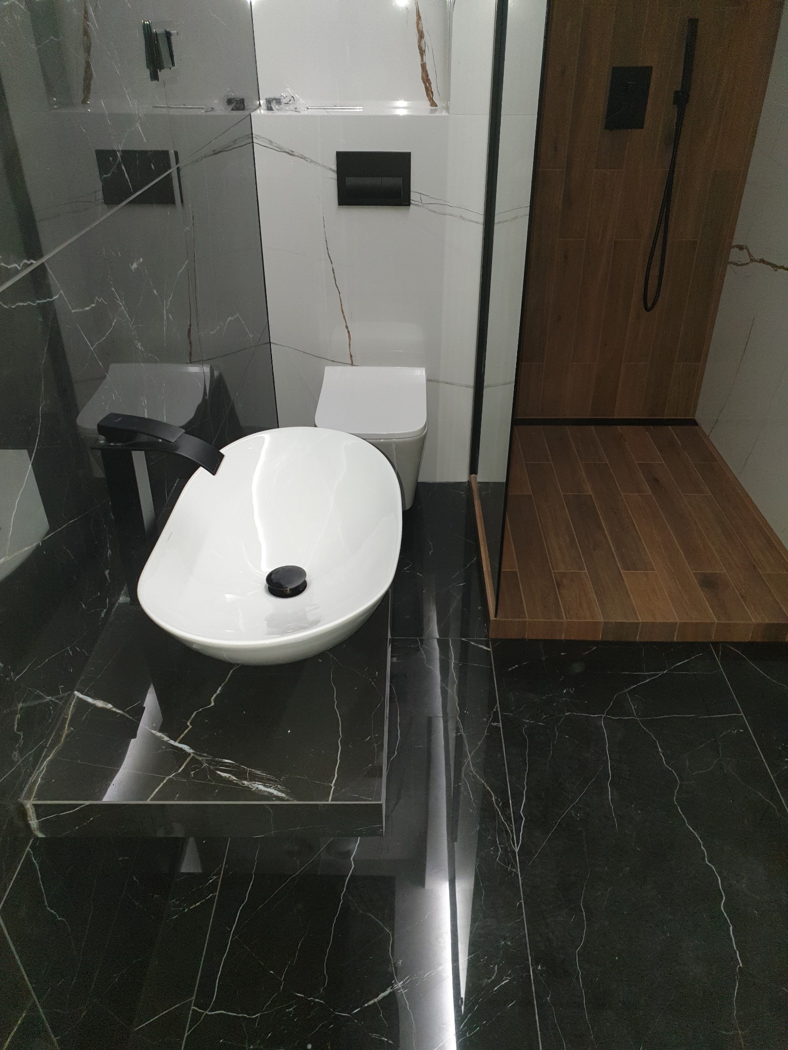 Nowoczesna łazienka z czarnymi marmurowymi płytkami, białą umywalką nablatową z czarną armaturą, toaletą podwieszaną i kabiną prysznicową typu walk-in z drewnopodobną podłogą.