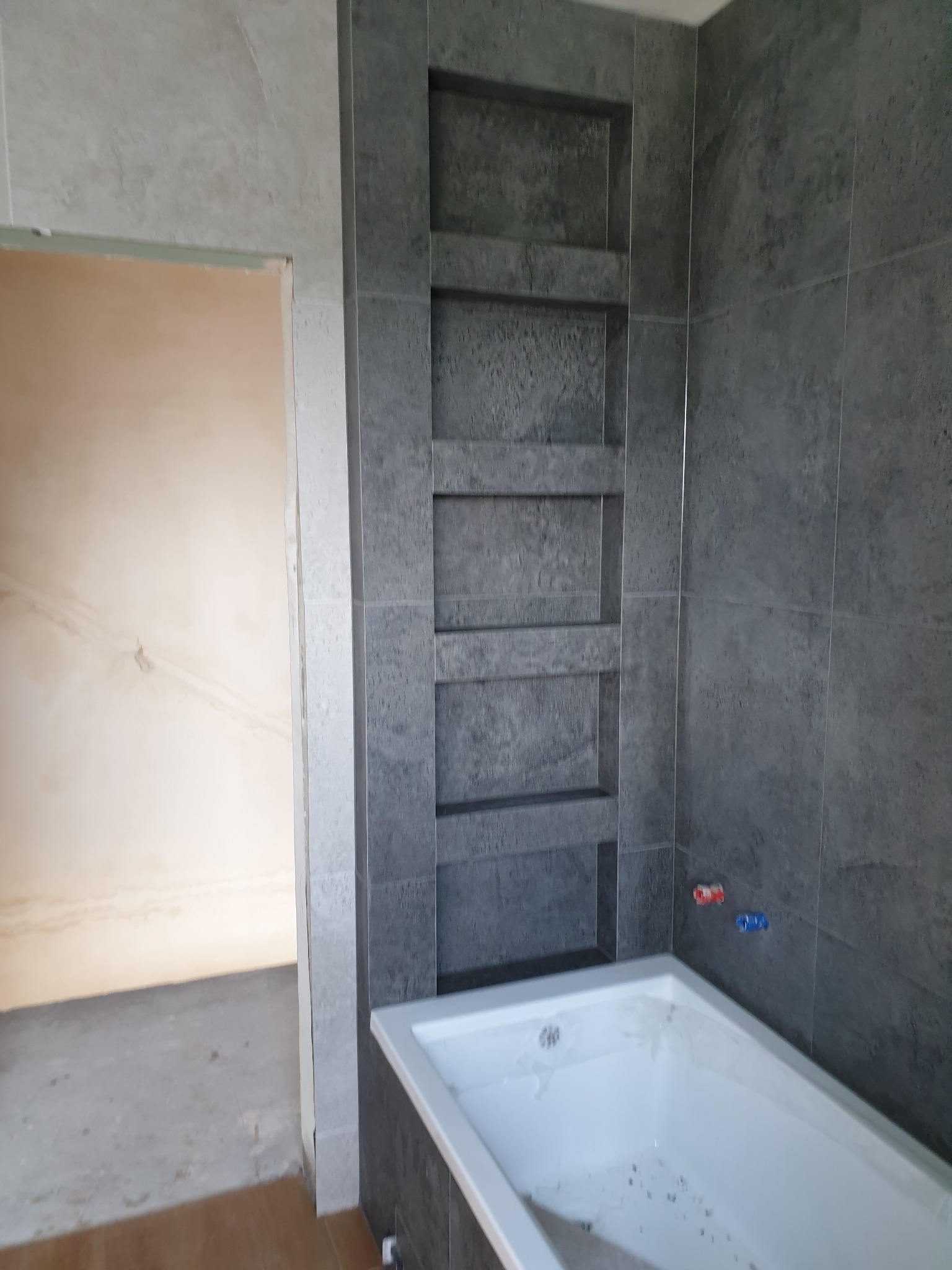 Łazienka w trakcie remontu: szare płytki z wnękami, biała wanna, widoczne przyłącza wodne i surowa ściana w sąsiednim pomieszczeniu.