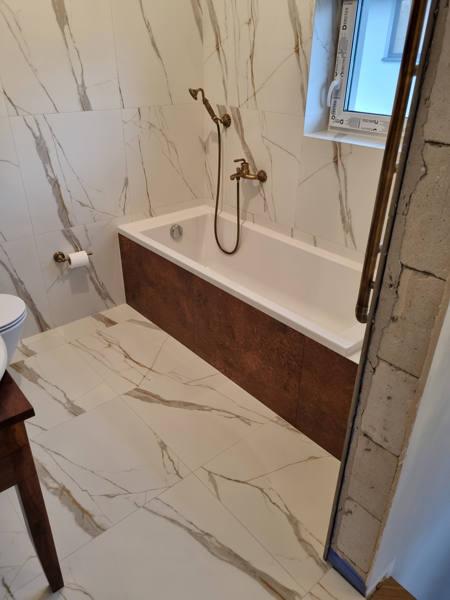Biała wanna z brązowym panelem obudowy, armatura w stylu retro, płytki imitujące marmur na ścianach i podłodze, widoczna surowa ściana w trakcie prac wykończeniowych