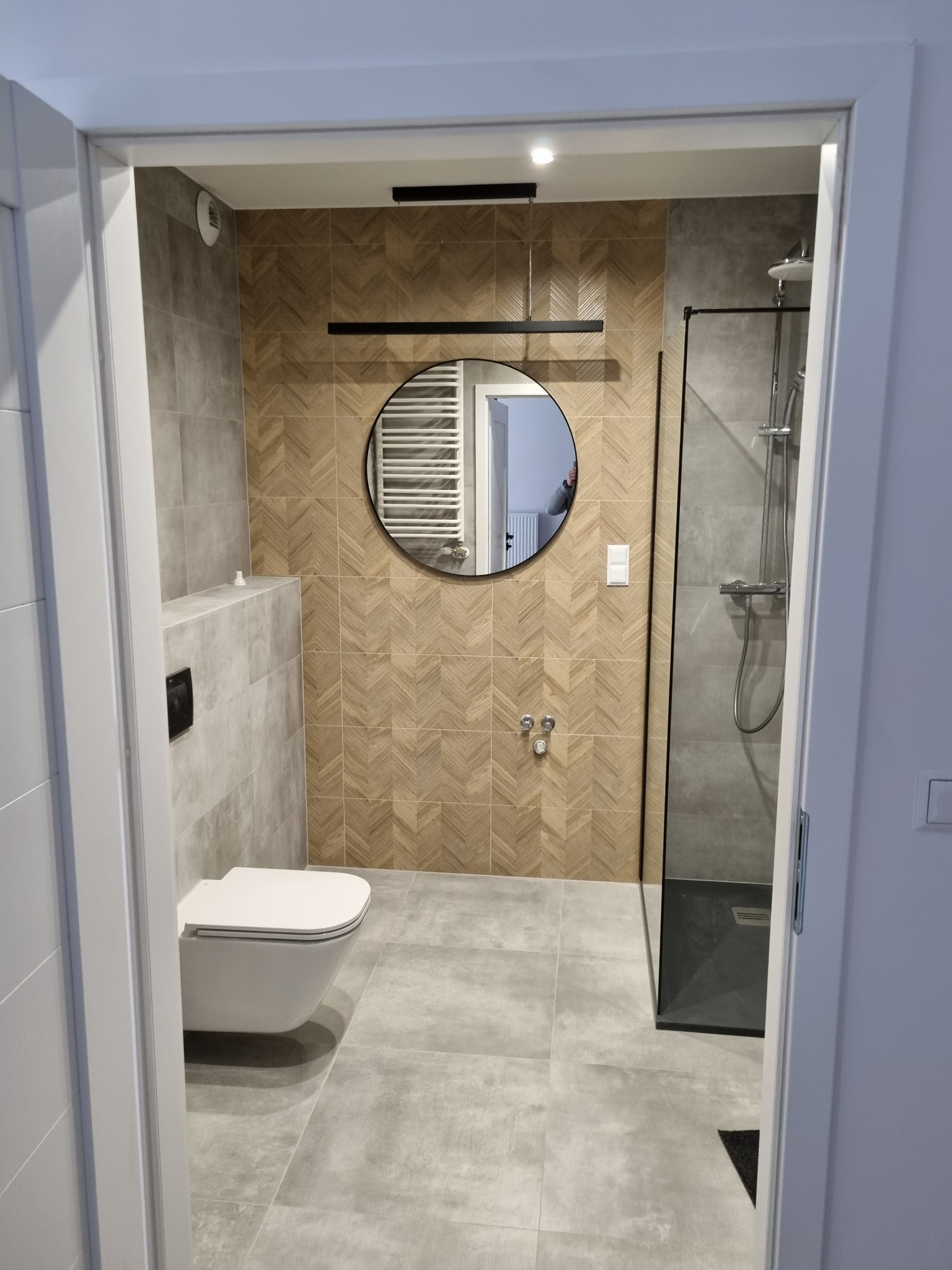 Nowoczesna łazienka z prysznicem typu walk-in, toaletą podwieszaną, okrągłym lustrem i płytkami imitującymi drewno ułożonymi w jodełkę na jednej ze ścian.