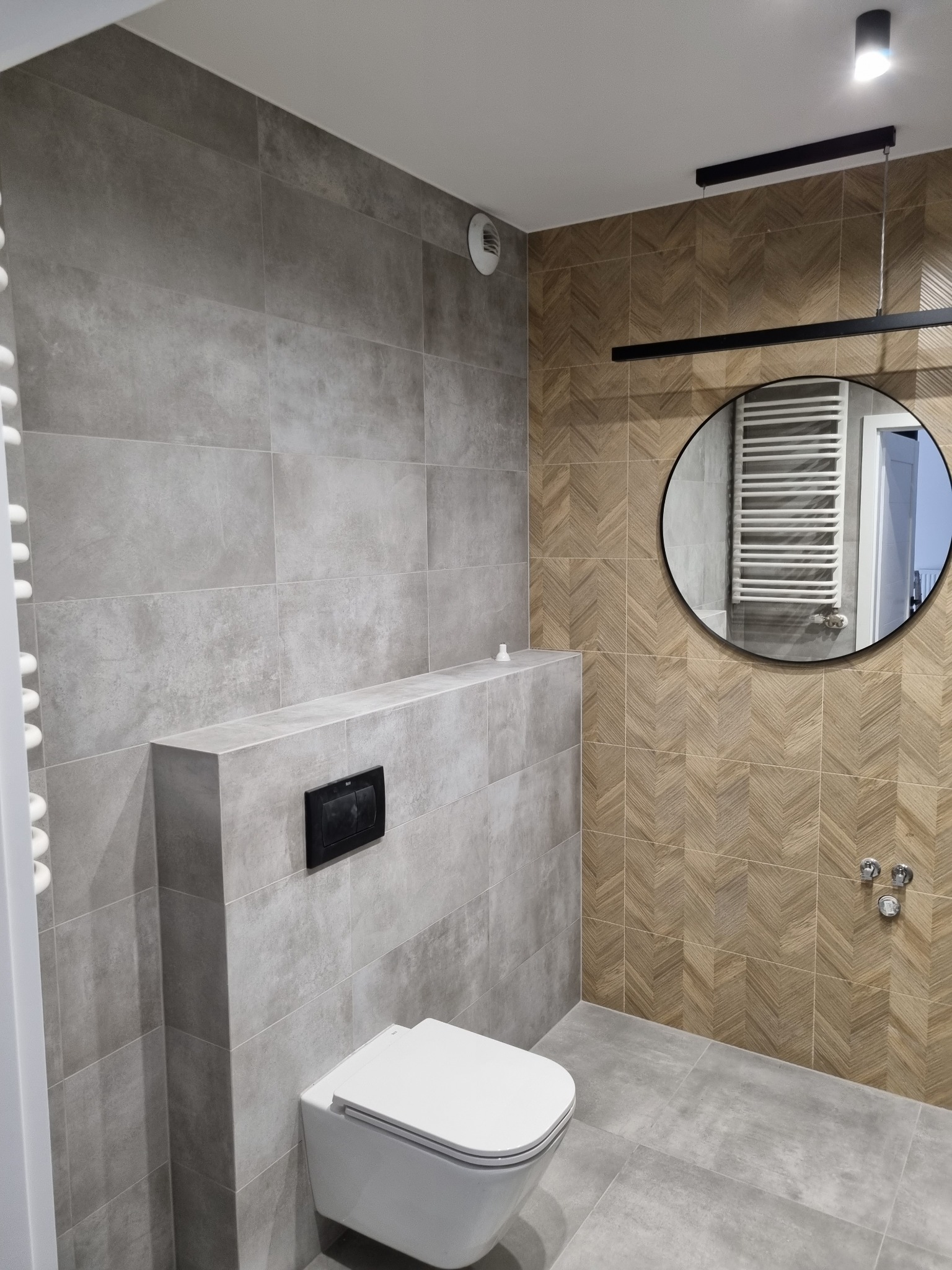 Nowoczesna łazienka z szarymi płytkami imitującymi beton, podwieszana toaleta z czarnym przyciskiem, ściana z drewnopodobnych płytek ułożonych w jodełkę, okrągłe lustro z czarną ramą i grzejnik...