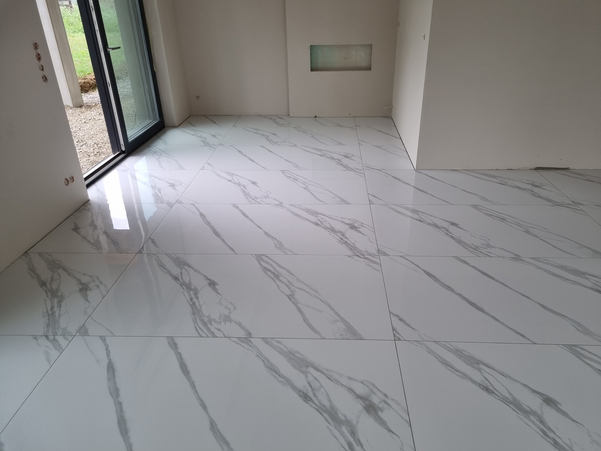 Błyszczące, białe płytki podłogowe imitujące marmur w nowym, jasnym pomieszczeniu z widocznym oknem i przygotowaną wnęką w ścianie.