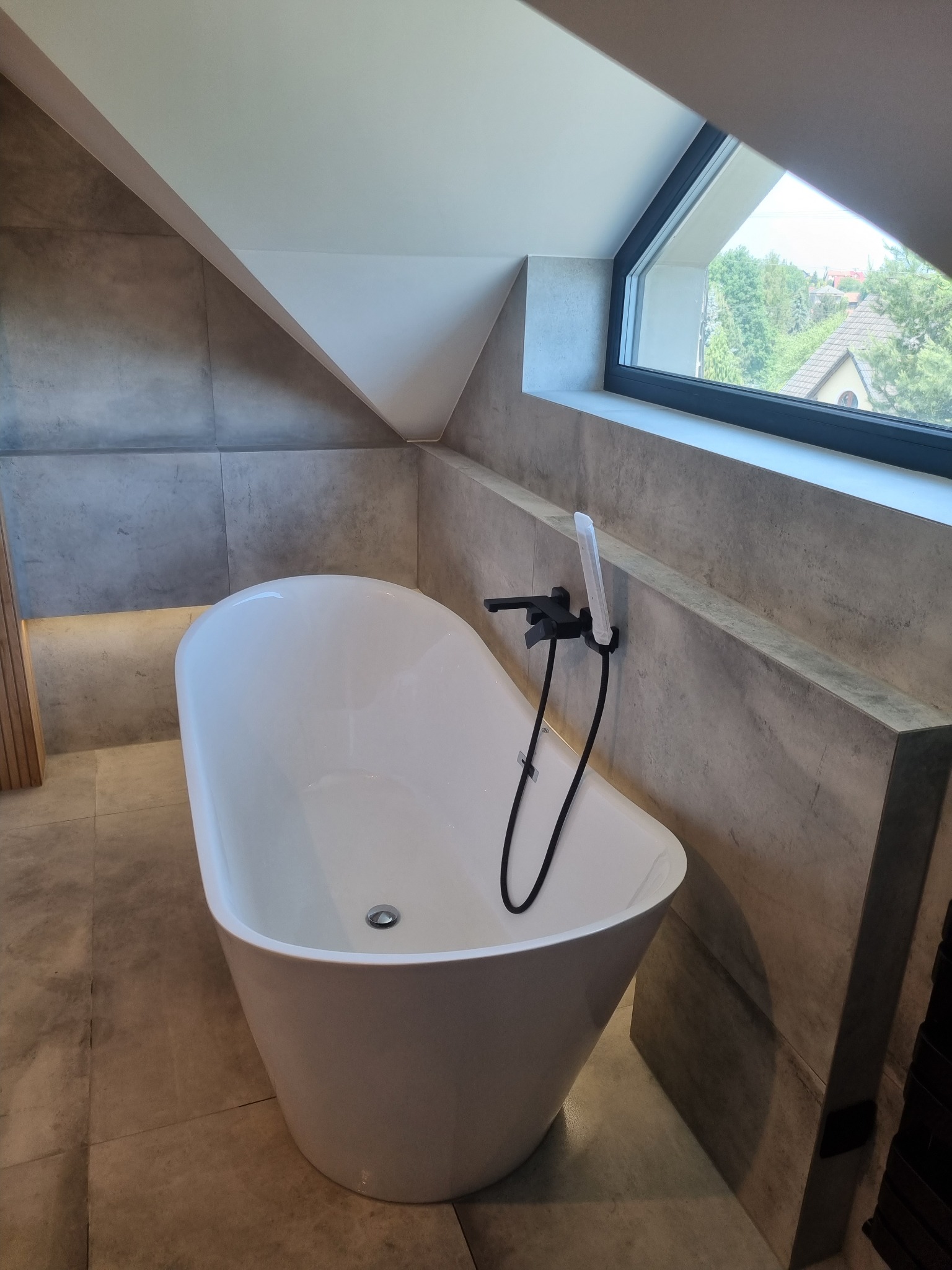 Wolnostojąca, biała wanna z czarną armaturą w łazience na poddaszu w Rzezawie. Okładzina ścian z płytek imitujących beton, okno dachowe z widokiem na zieleń.