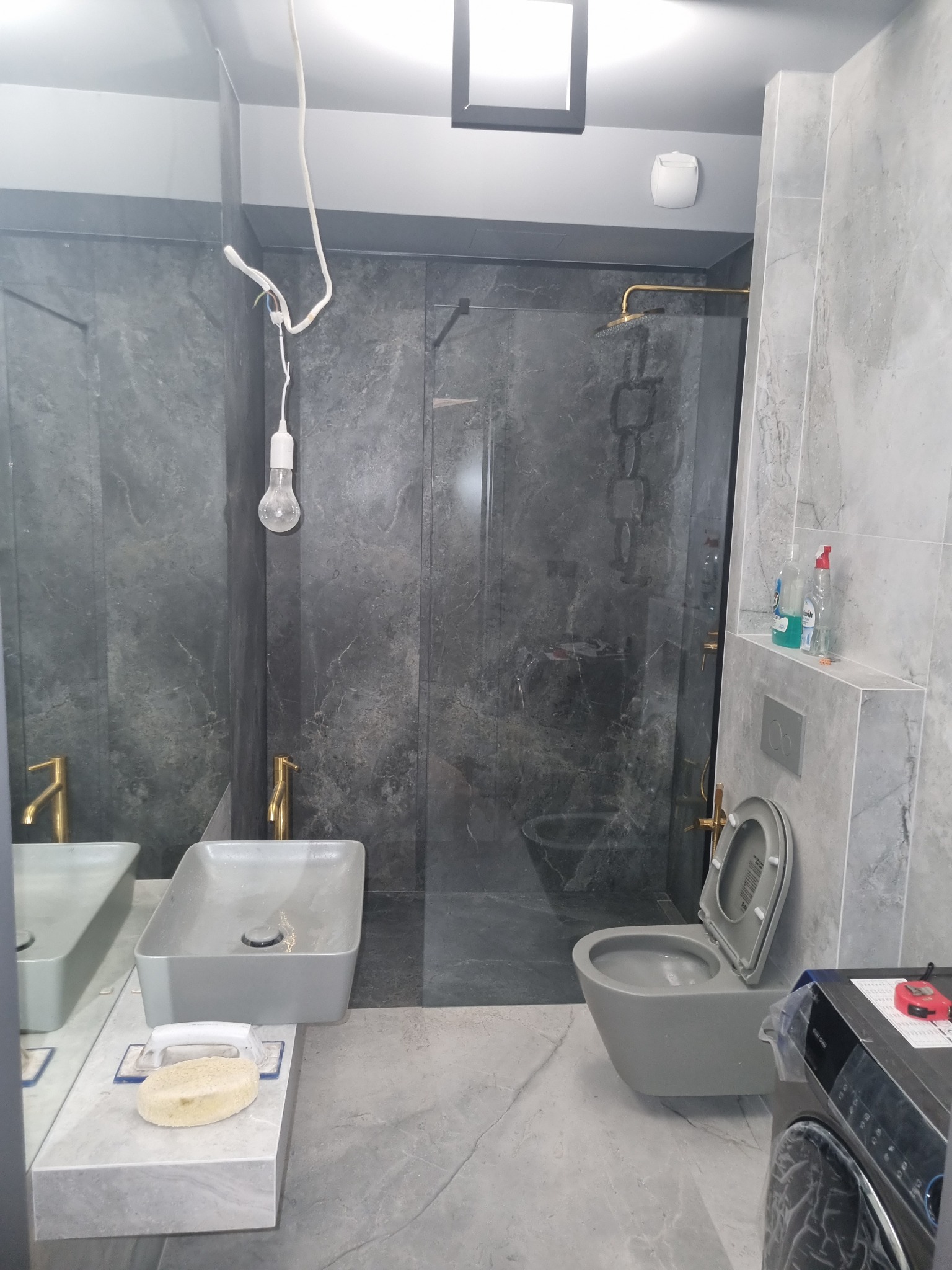 Szara łazienka z kabiną prysznicową, umywalką nablatową i podwieszaną toaletą w trakcie remontu, widoczne niepodłączone kable i żarówka.