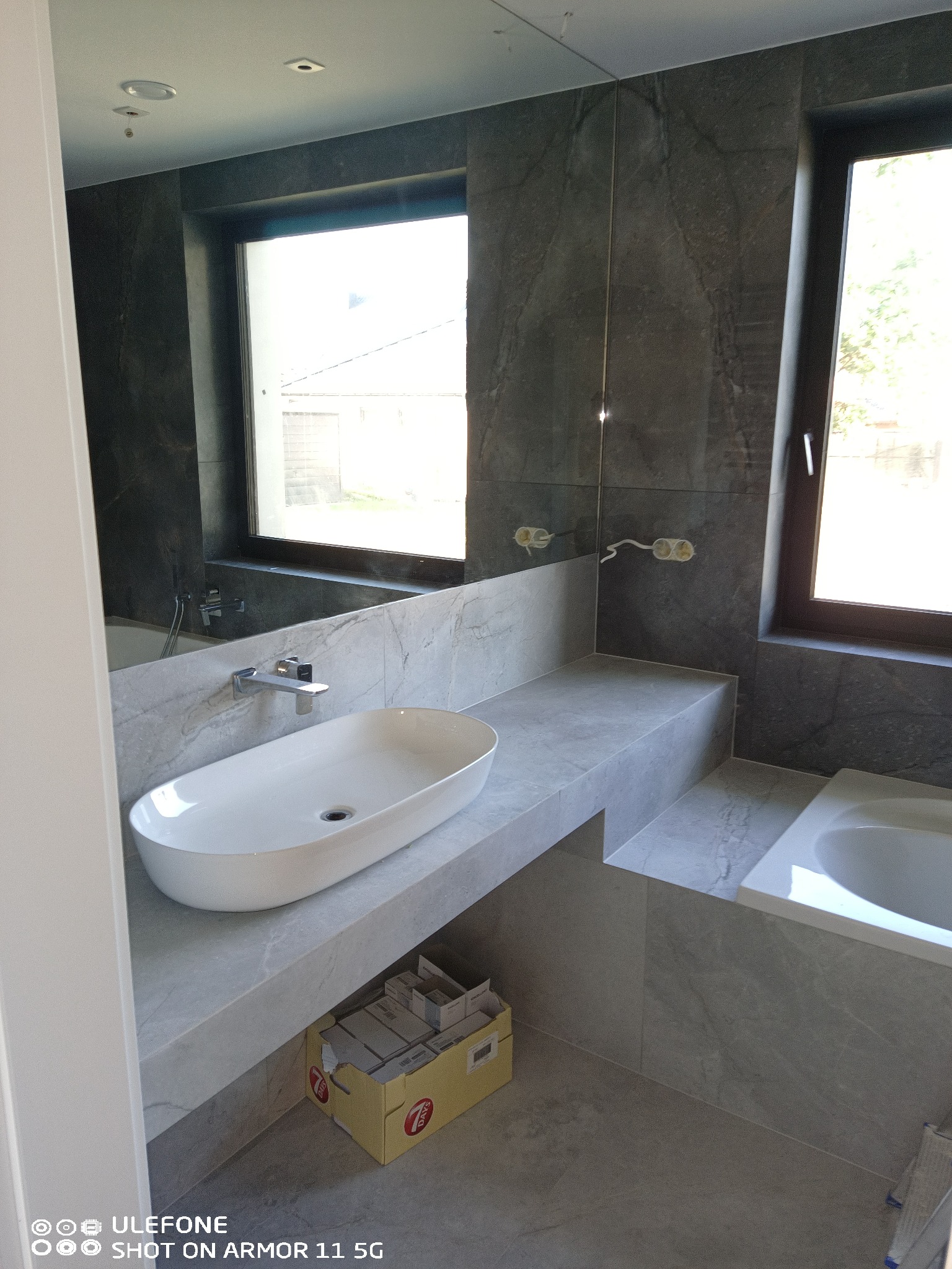 Nowoczesna łazienka w szarościach z owalną umywalką nablatową i dużym lustrem, widoczne przygotowane kable elektryczne i kartonowe opakowanie płytek pod umywalką.