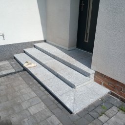 Łukasz Chudziński - Granitowe schody wejściowe do domu z beżowymi klapkami leżącymi na drugim stopniu, obok widoczny chodnik z kostki brukowej i fragment ściany z kranem.