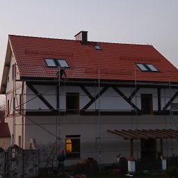 Łukasz Chudziński - Praca na wysokości: dekarz na rusztowaniu montuje czerwoną dachówkę na dachu domu z oknami dachowymi, widoczne cegły na ziemi.