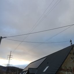 Łukasz Chudziński - Ciemnoszary dach z panelami słonecznymi i oknami dachowymi na tle pochmurnego nieba i linii energetycznych.