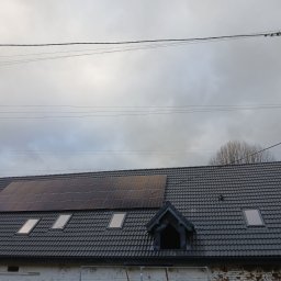 Łukasz Chudziński - Szary dach z dachówki z oknami dachowymi i panelami słonecznymi, widok z dołu na tle pochmurnego nieba i słupa energetycznego.