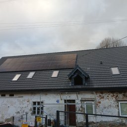 Łukasz Chudziński - Dom z szarą dachówką, panelami słonecznymi i oknami dachowymi, elewacja częściowo odnowiona, widoczne rusztowania i tablice informacyjne.
