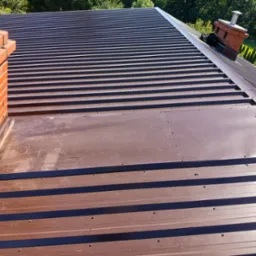 Nowe, brązowe pokrycie dachu z blachy trapezowej, widoczne kominy z cegły. Dach połaciowy z wyraźnymi liniami, świeżo po montażu, na tle zieleni drzew.