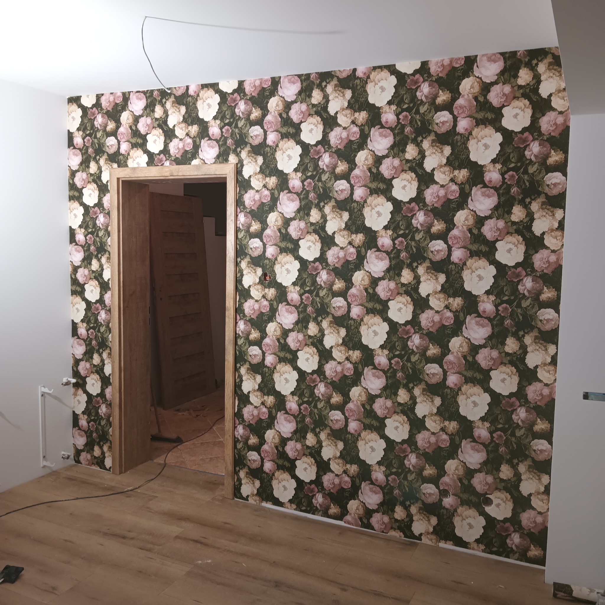 Pokój po remoncie z nową tapetą w kwiaty i odnowioną drewnianą futryną drzwiową, widoczna podłoga w panelach.