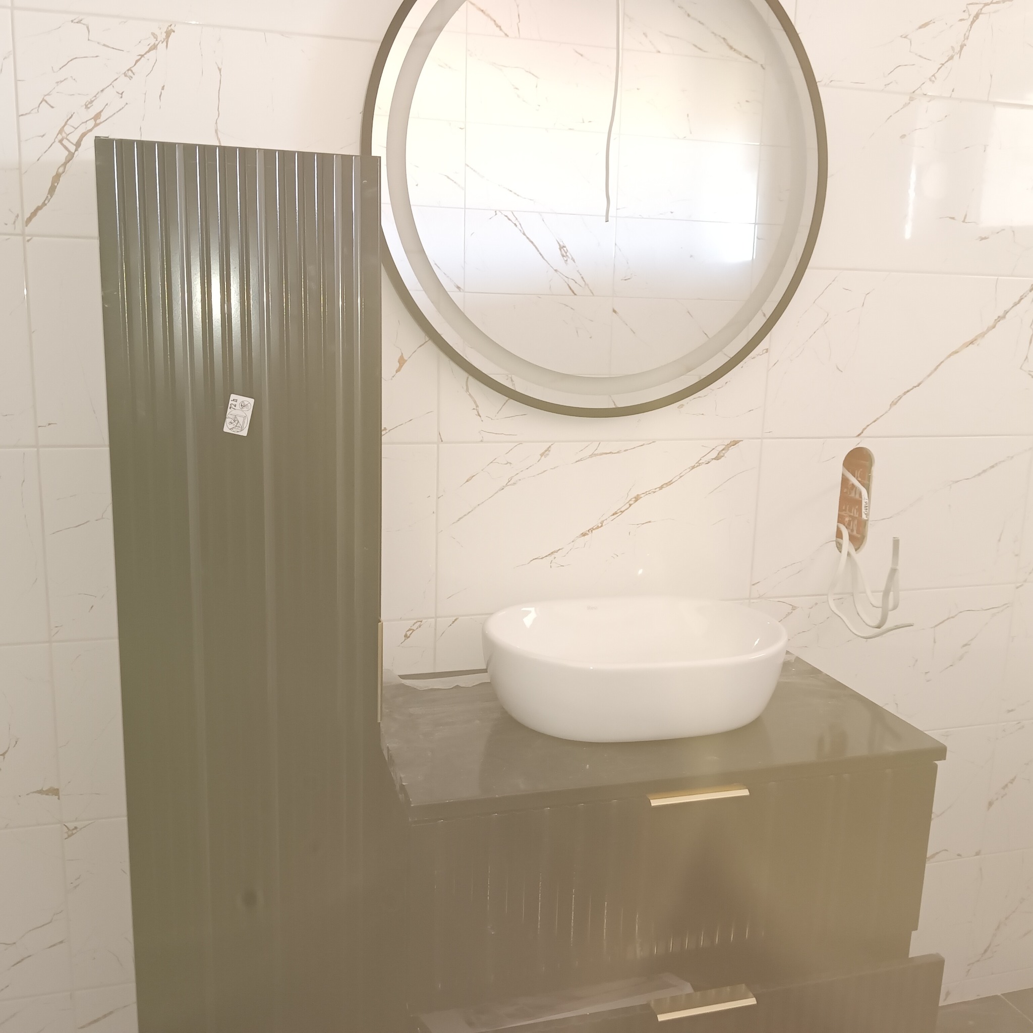 Nowoczesna umywalka nablatowa w łazience z marmurowymi płytkami, okrągłe lustro z podświetleniem, szara szafka z frezowanymi frontami i złotymi uchwytami, widoczne akcesoria łazienkowe.