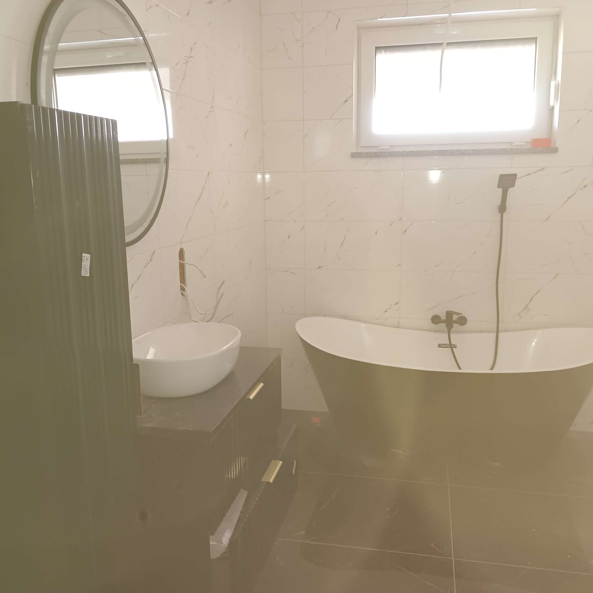 Nowoczesna łazienka z wolnostojącą wanną w kolorze oliwkowym, białą umywalką nablatową i szafką w ciemnym odcieniu zieleni, marmurowe płytki na ścianach i podłodze, okrągłe lustro i okno z widokiem...