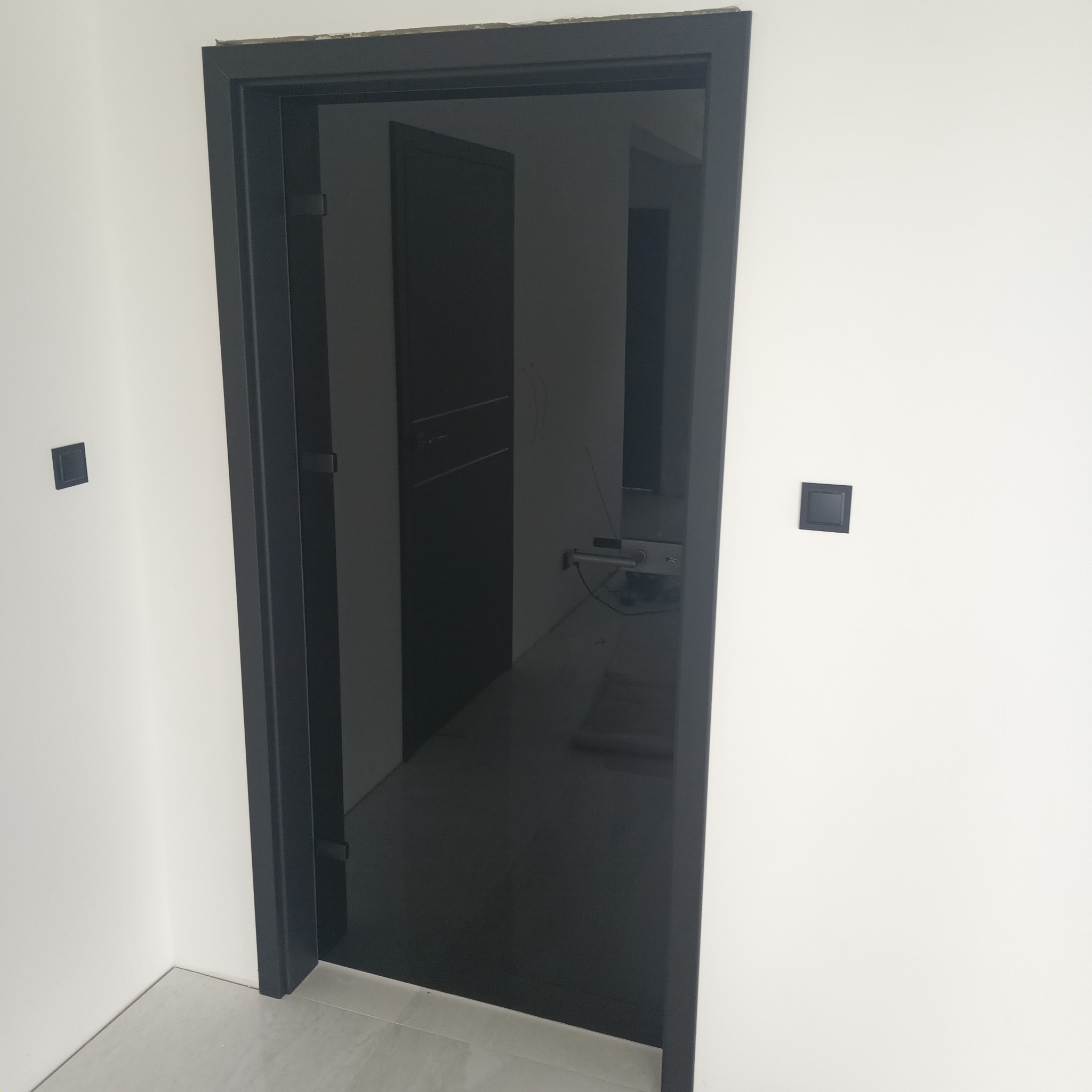 Czarne, minimalistyczne drzwi wewnętrzne z widocznym fragmentem kolejnych drzwi i czarnymi włącznikami na białej ścianie.