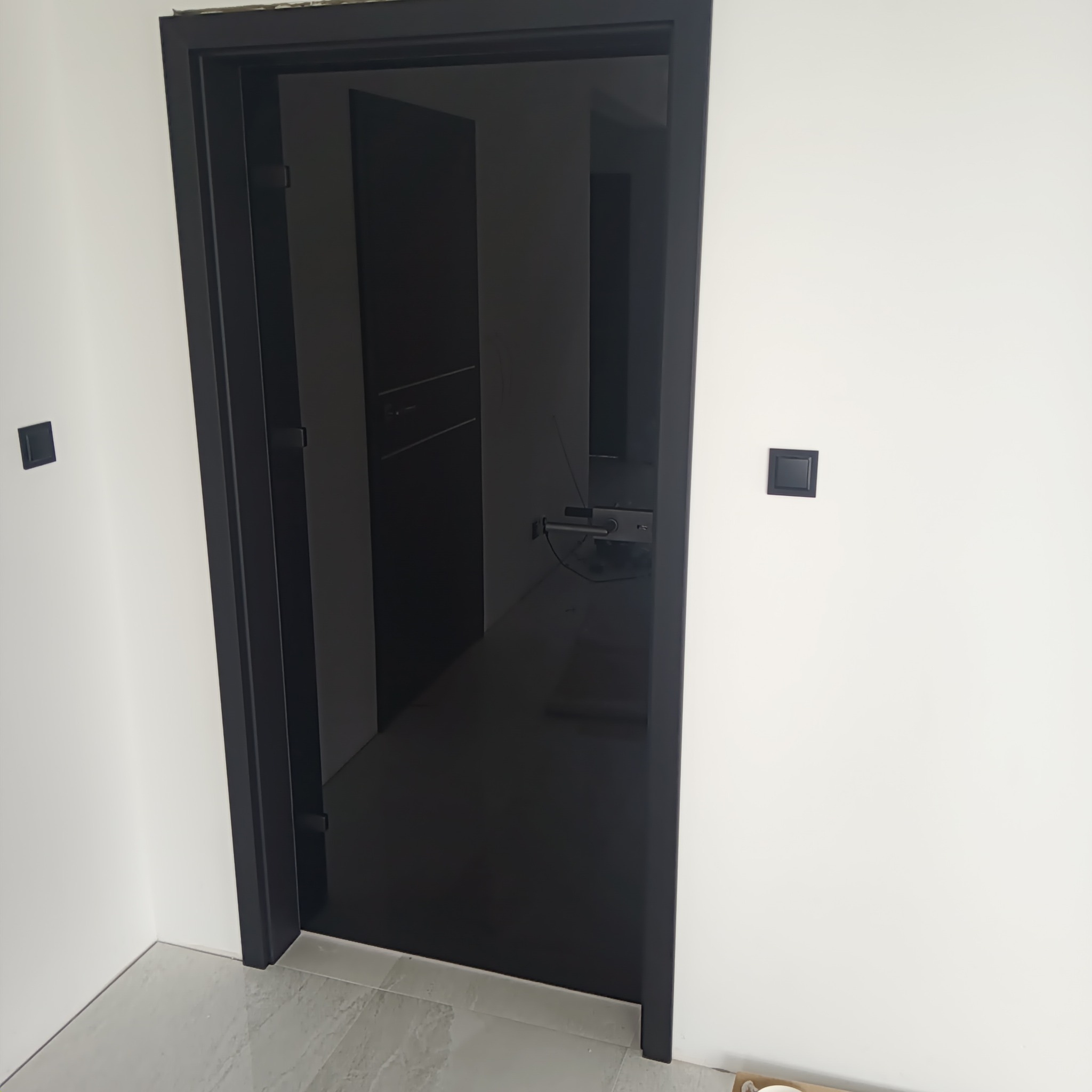 Zamontowane, nowoczesne, czarne drzwi wewnętrzne z widocznym fragmentem pomieszczenia za nimi, białe ściany z czarnymi włącznikami, jasna podłoga.