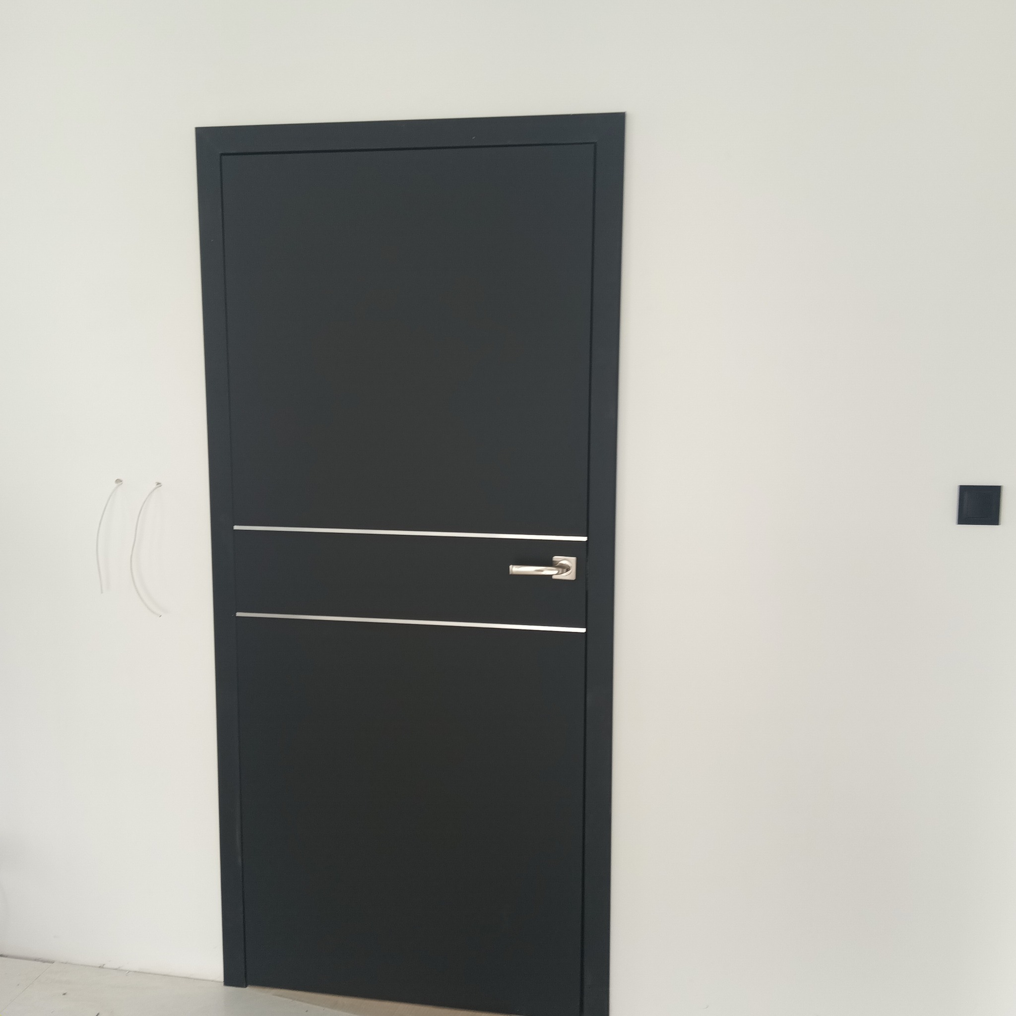 Ciemne, nowoczesne drzwi wewnętrzne z srebrnymi akcentami i klamką, zamontowane w białej ścianie. Widoczne wystające kable po lewej stronie.