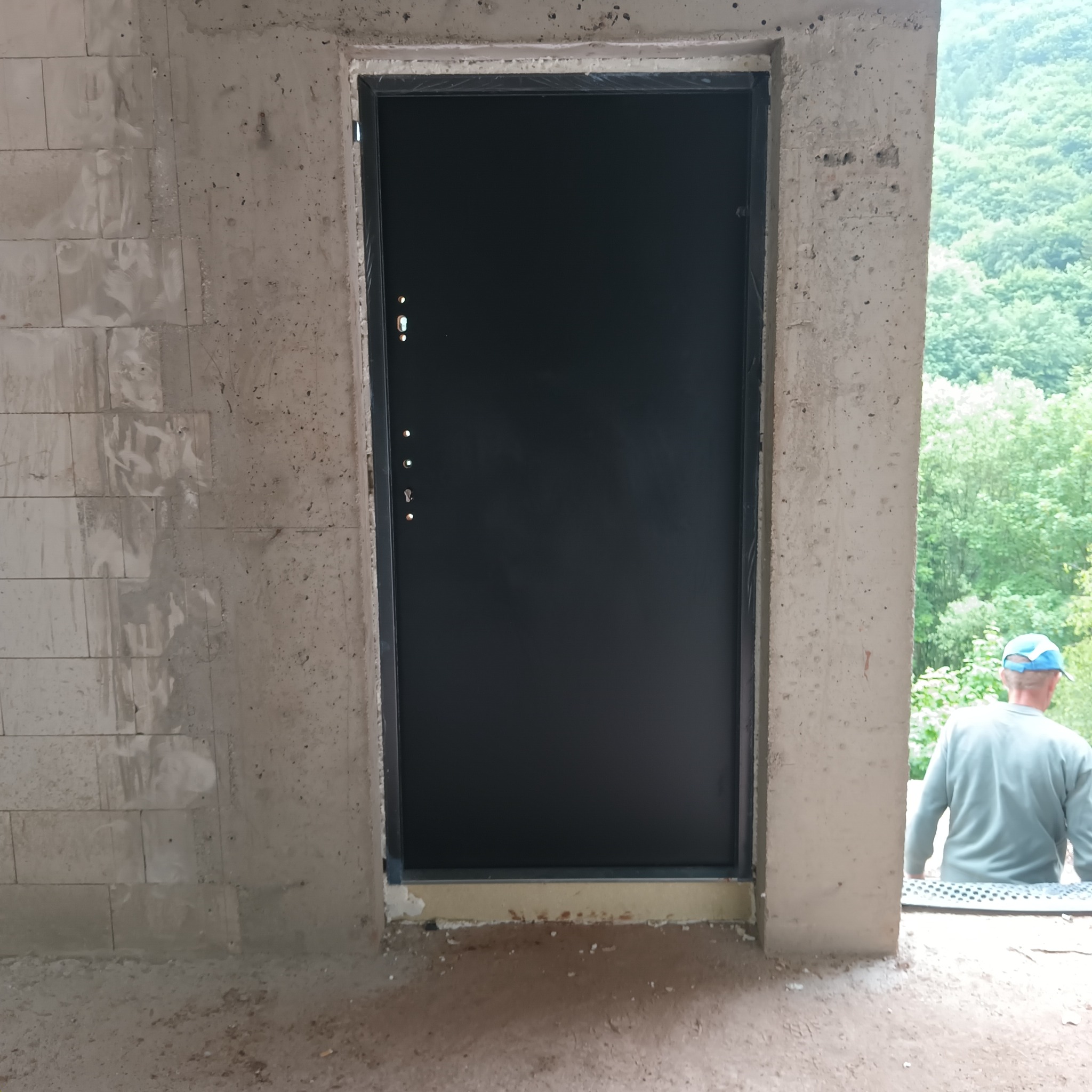 Czarna, antywłamaniowa brama w surowym otoczeniu betonowych ścian i podłogi, z widocznym pracownikiem w tle i zielenią drzew za oknem.