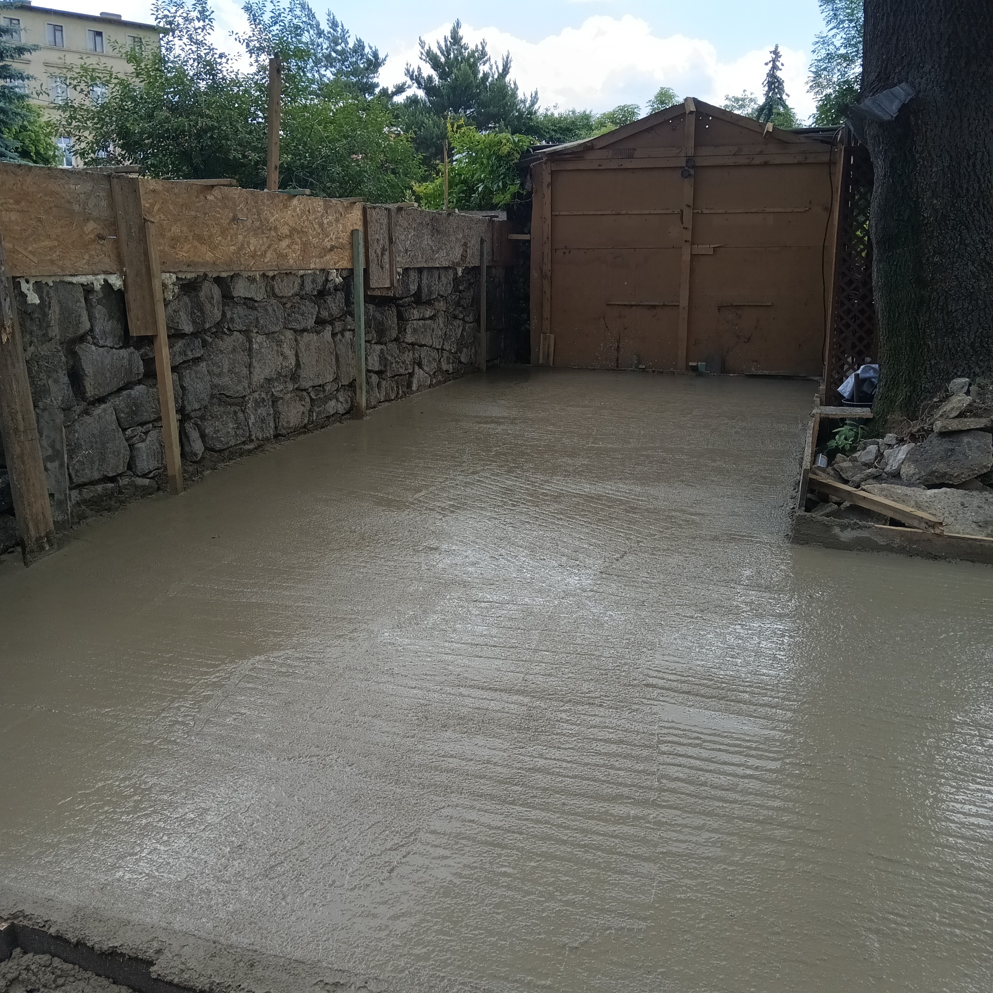 Świeżo wylana, mokra posadzka betonowa na zewnątrz, z widocznym szalunkiem i kamiennym murem w tle, pod drewnianym garażem.