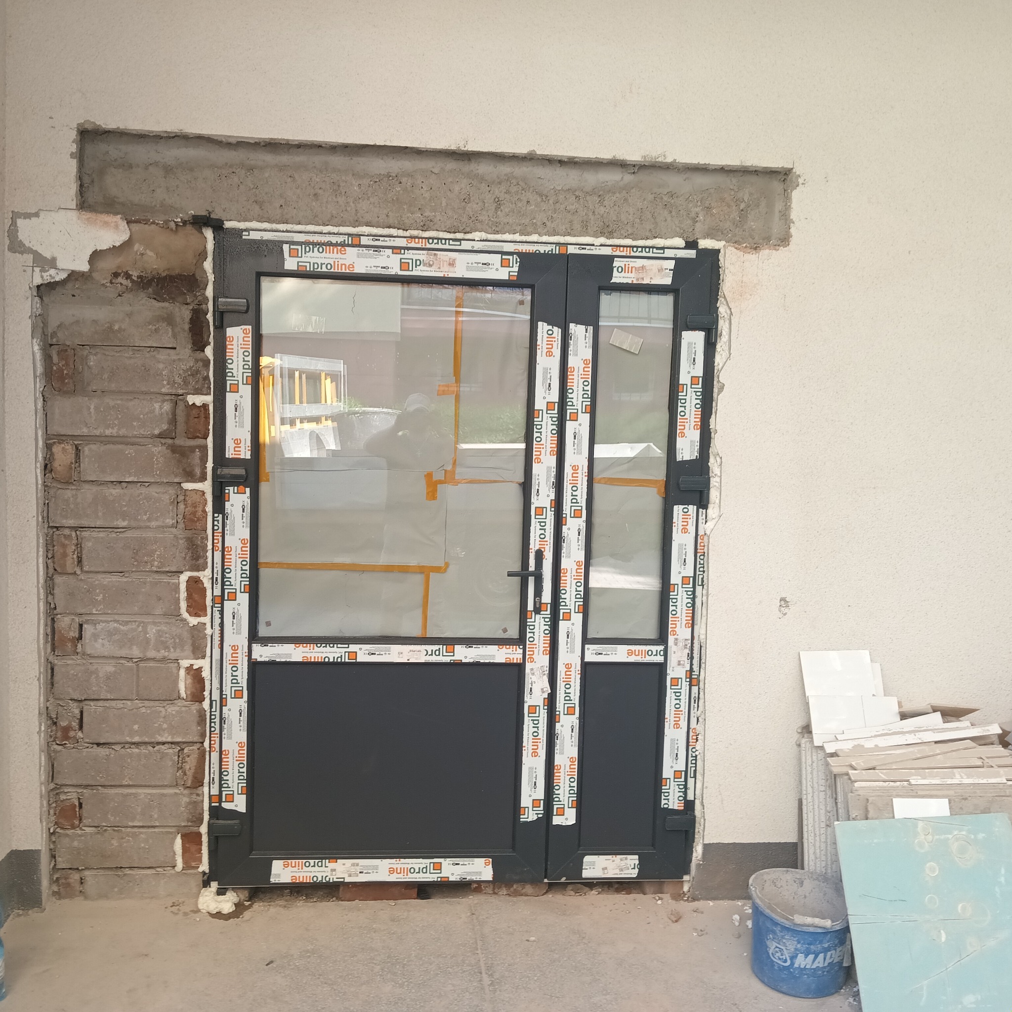 Ciemne, dwuskrzydłowe drzwi balkonowe z szybami, zabezpieczone folią, w trakcie montażu w surowym otworze ściennym z widoczną cegłą i pianką montażową. Na ramie drzwi naklejone taśmy z logo...