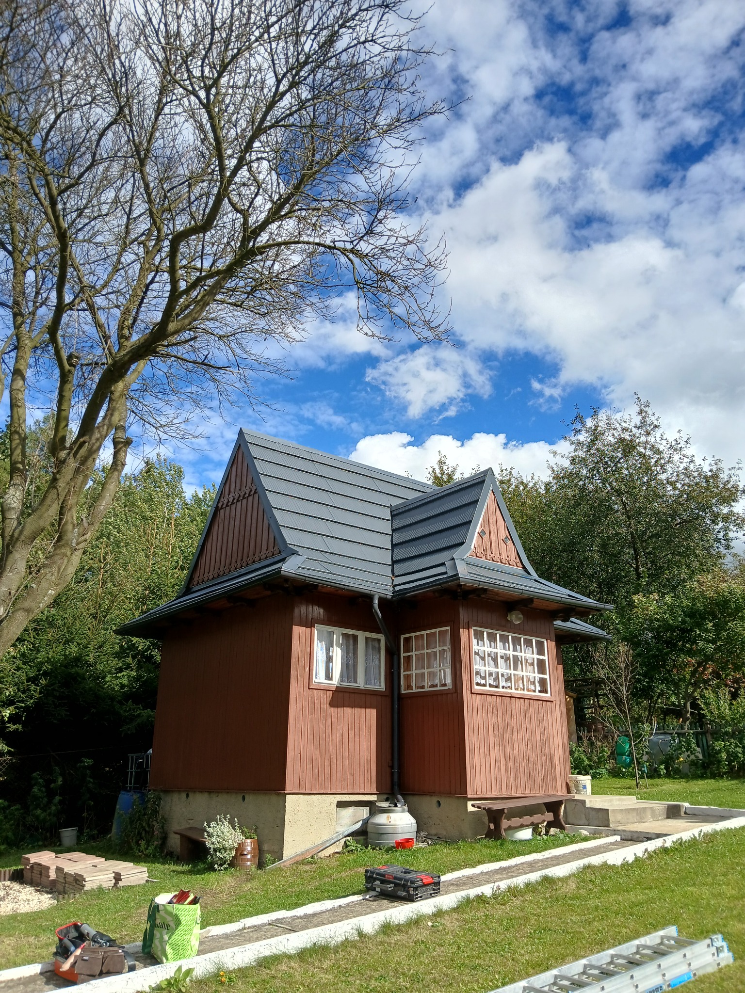 Drewniany domek z nowym, grafitowym dachem z blachodachówki modułowej, widok z poziomu gruntu, na trawniku leżą narzędzia i materiały budowlane.