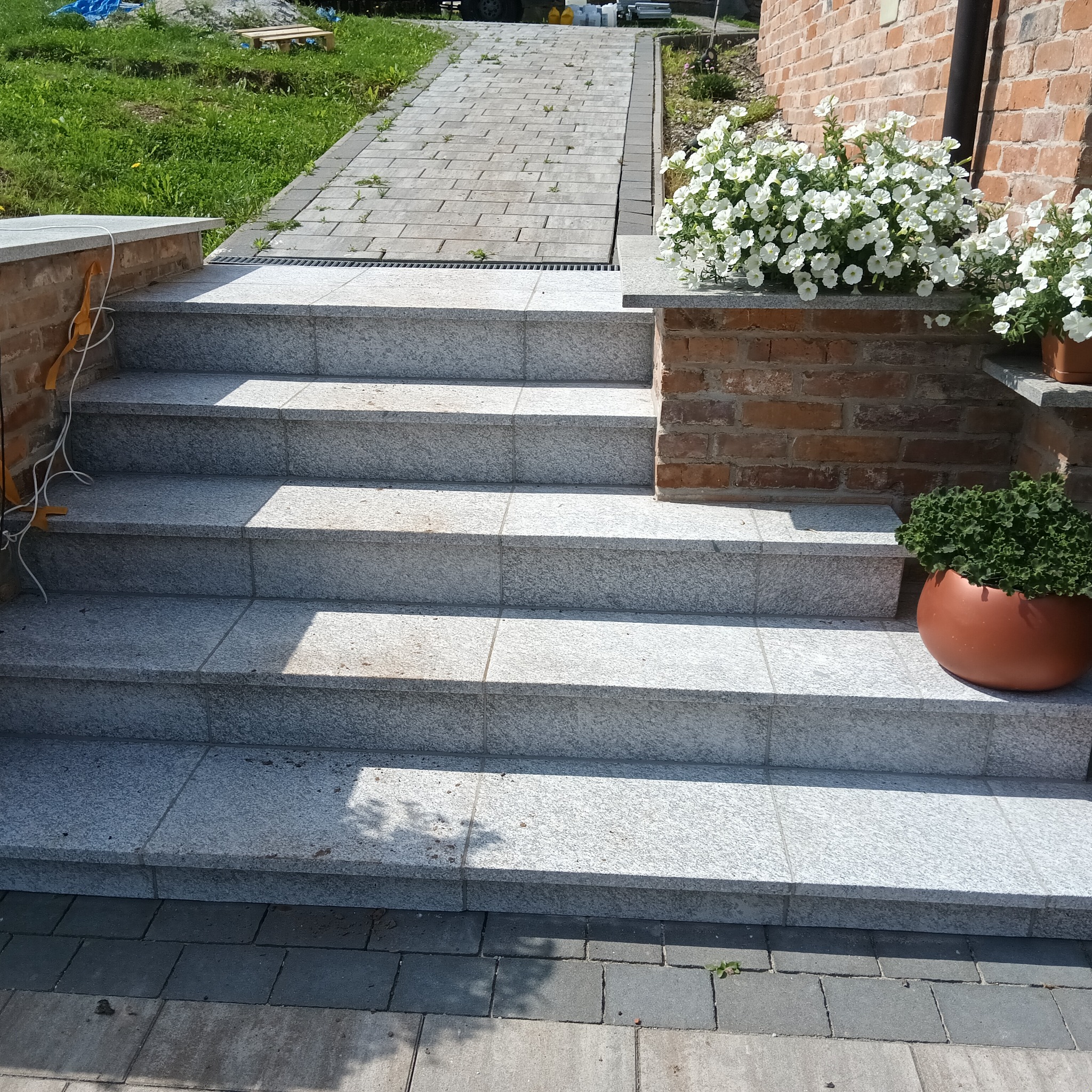 Granitowe schody zewnętrzne prowadzące do domu z cegły, ozdobione białymi kwiatami w donicach i rośliną w brązowej donicy, z widoczną ścieżką z kostki brukowej.