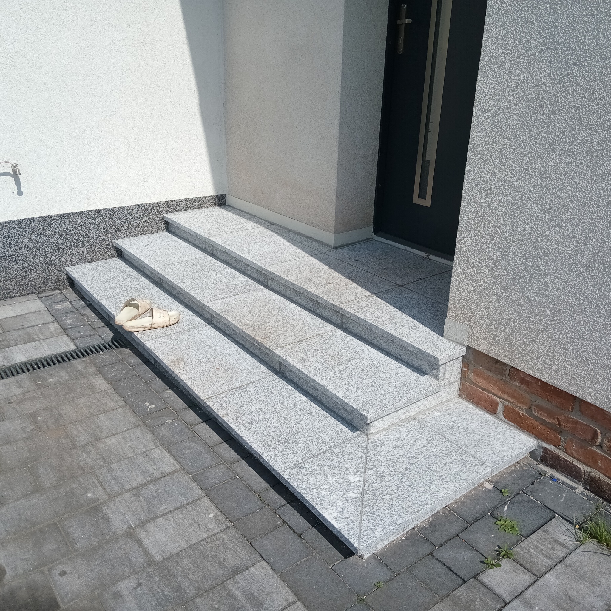 Granitowe schody wejściowe do domu z beżowymi klapkami leżącymi na drugim stopniu, obok widoczny chodnik z kostki brukowej i fragment ściany z kranem.