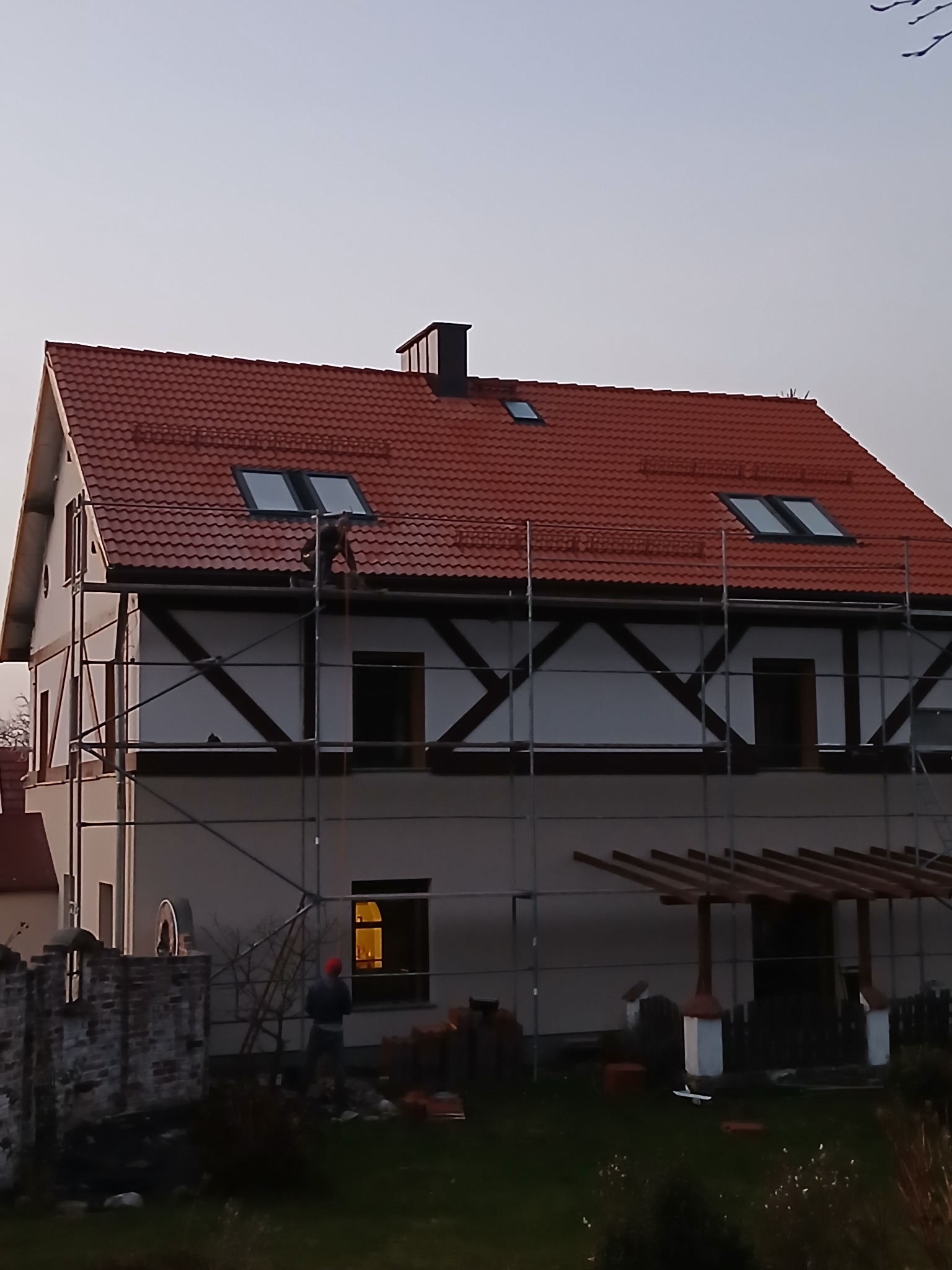 Praca na wysokości: dekarz na rusztowaniu montuje czerwoną dachówkę na dachu domu z oknami dachowymi, widoczne cegły na ziemi.