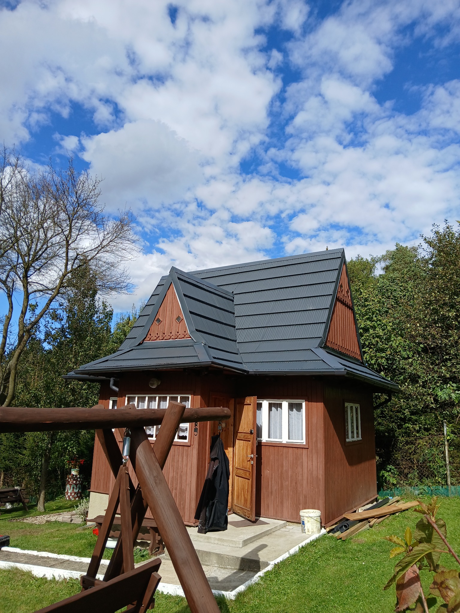 Chatka z nowym, ciemnoszarym dachem w stylu góralskim, drewniana konstrukcja, błękitne niebo z chmurami w tle, huśtawka na pierwszym planie.