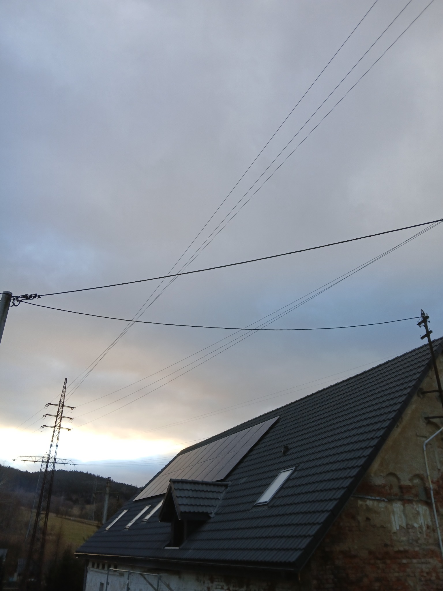 Ciemnoszary dach z panelami słonecznymi i oknami dachowymi na tle pochmurnego nieba i linii energetycznych.