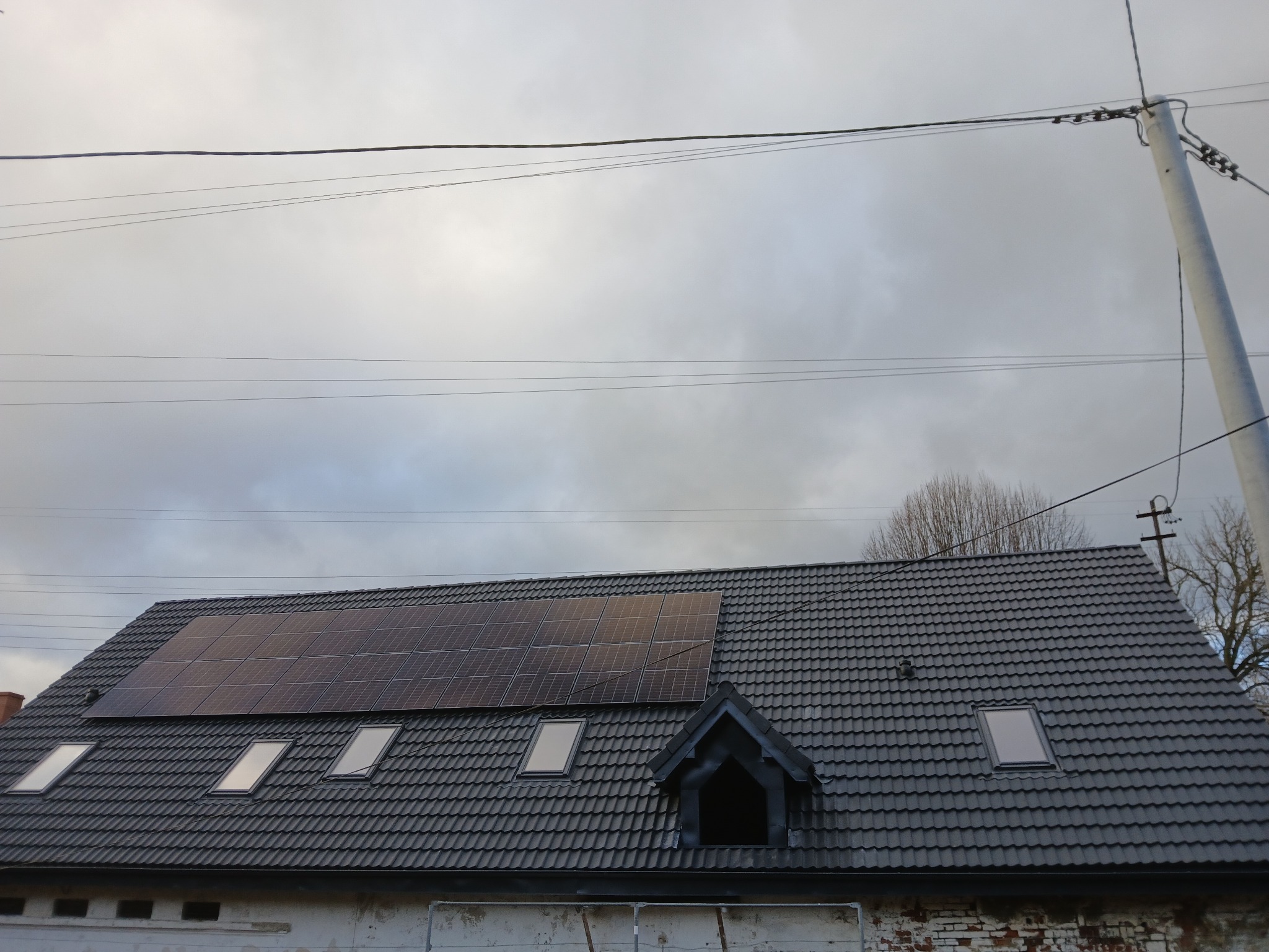 Szary dach z dachówki z oknami dachowymi i panelami słonecznymi, widok z dołu na tle pochmurnego nieba i słupa energetycznego.